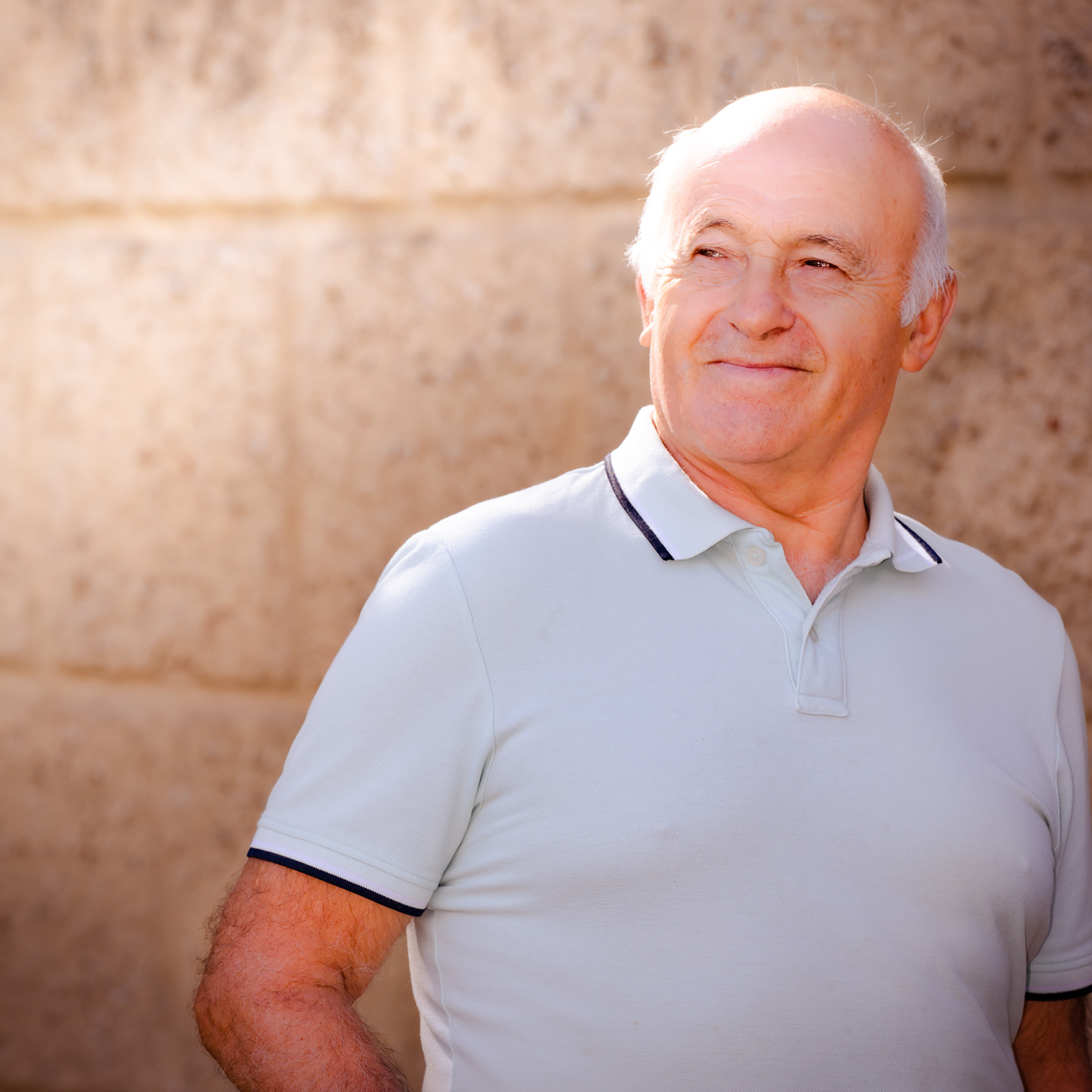 Ein lebendiges Porträt eines älteren, fast kahlköpfigen Mannes vor einer Steinmauer bei Sonnenuntergang.