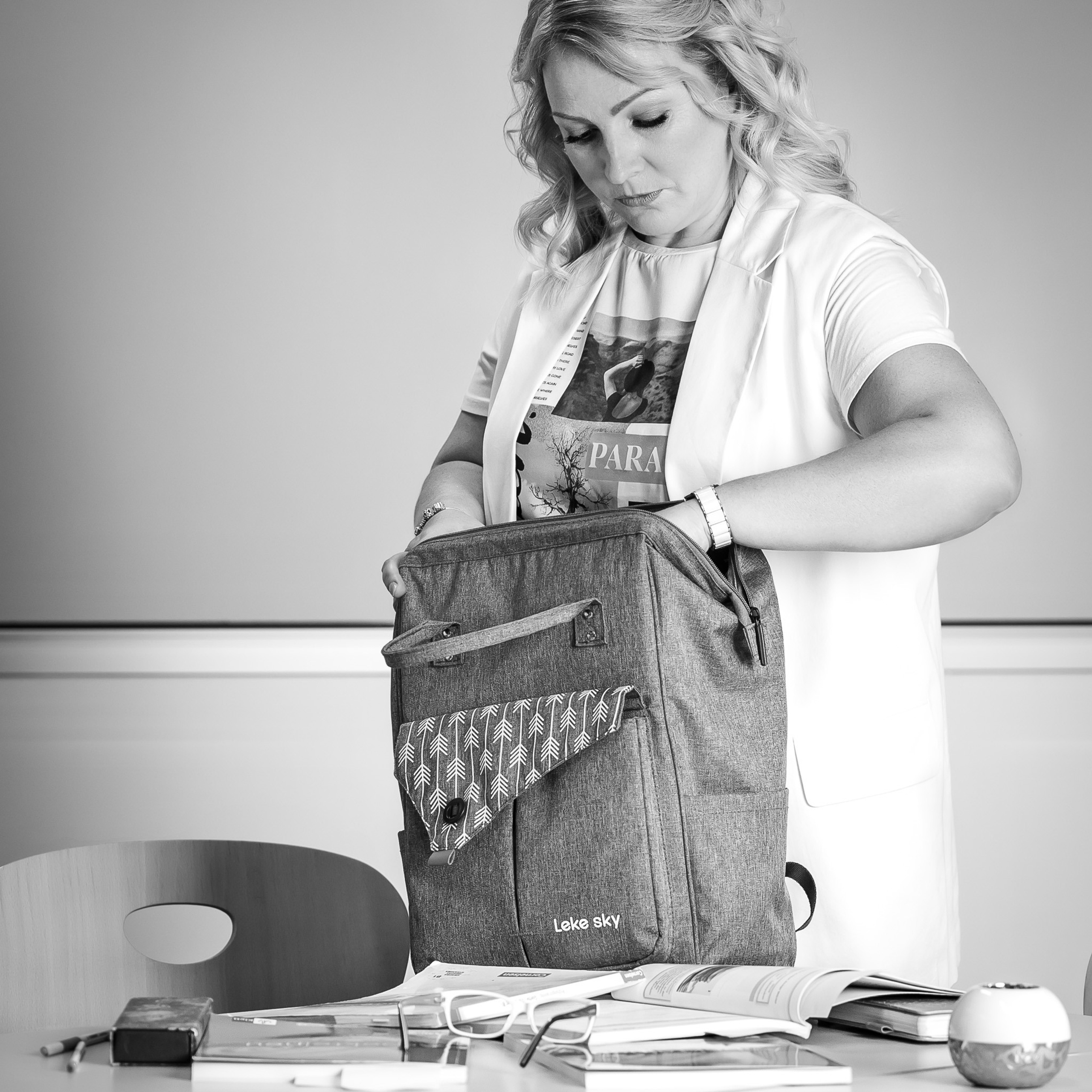 Ein schwarz-weißes Foto einer Studentin, die am Tisch mit Büchern steht und ihren Laptop aus ihrem Rucksack hervorholt.