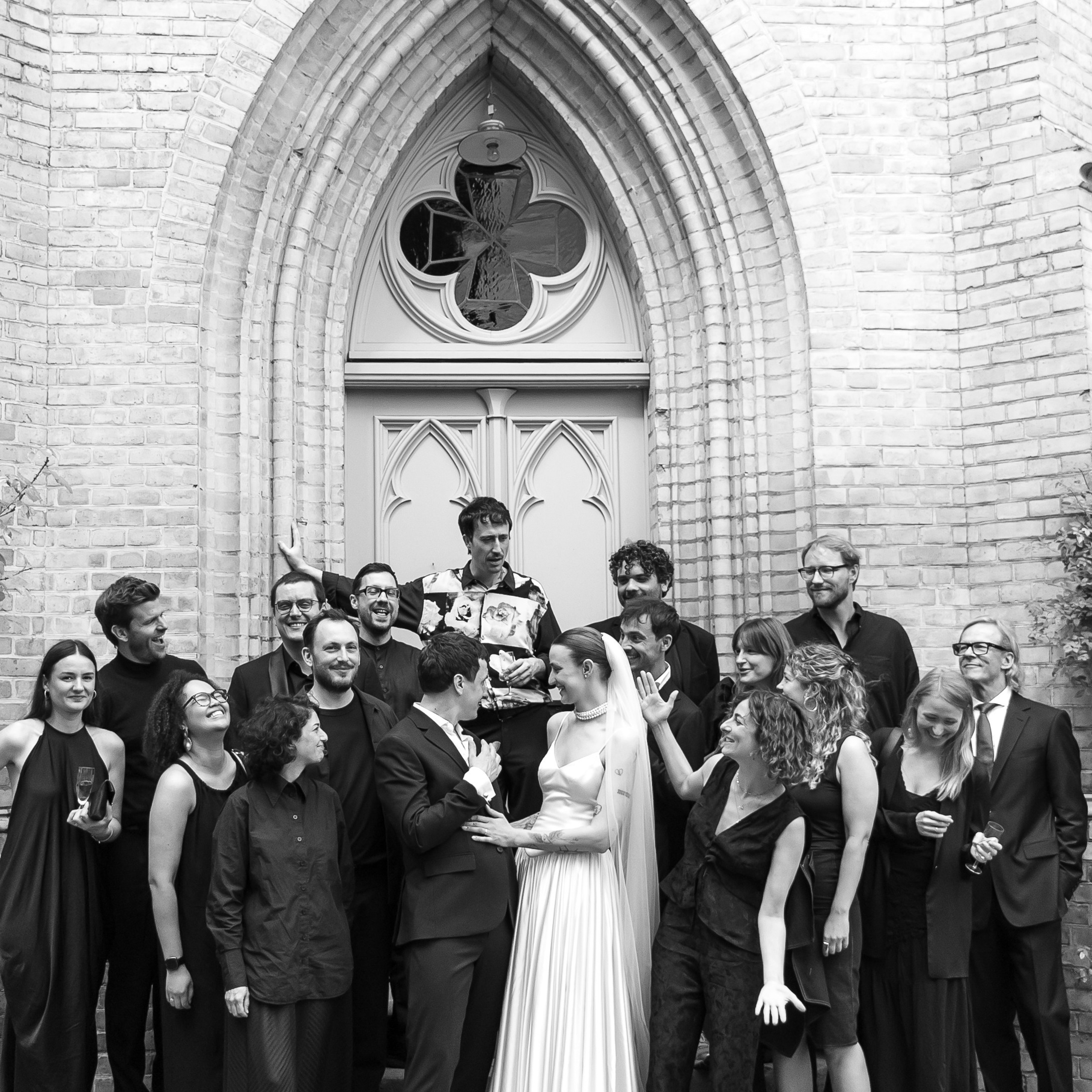 Schwarz-weißes Gruppenfoto von Brautpaar und Hochzeitsgästen vor dem gotischen Portal der Alten Neuendorfer Kirche in Potsdam