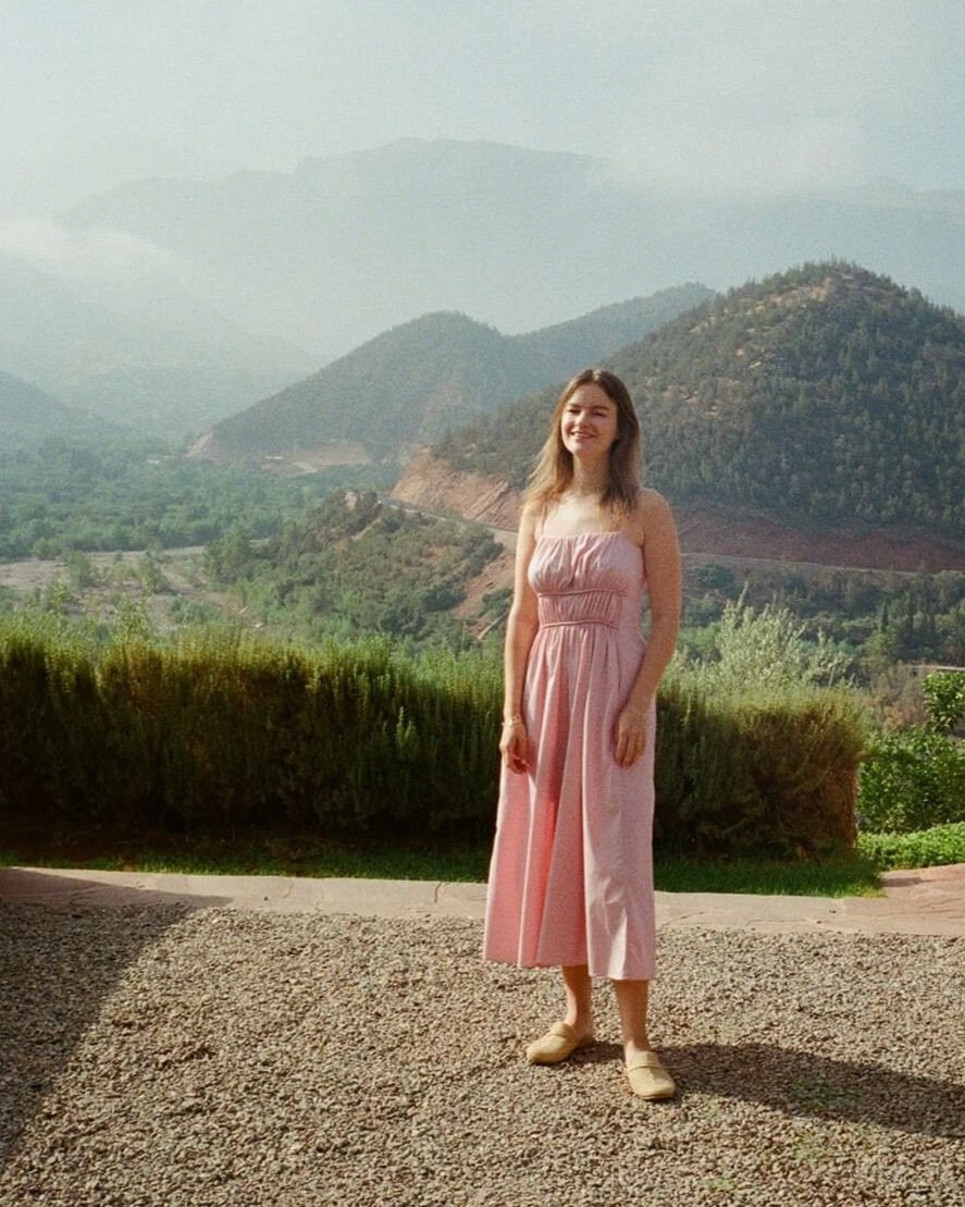 Destination wedding photographer Inna Zaytseva standing in a pink dress on a sunlit gravel terrace with layered mountain landscape behind