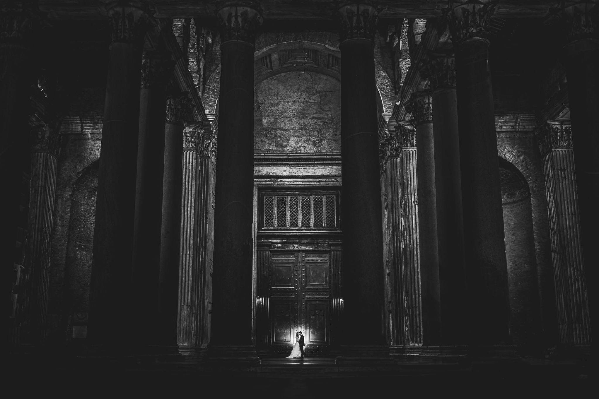 Coppia di sposi si tiene per mano al centro di un'antica architettura con colonne imponenti, illuminati da una singola fonte di luce in una maestosa hall al Pantheon creando un'atmosfera intima e storica in bianco e nero.