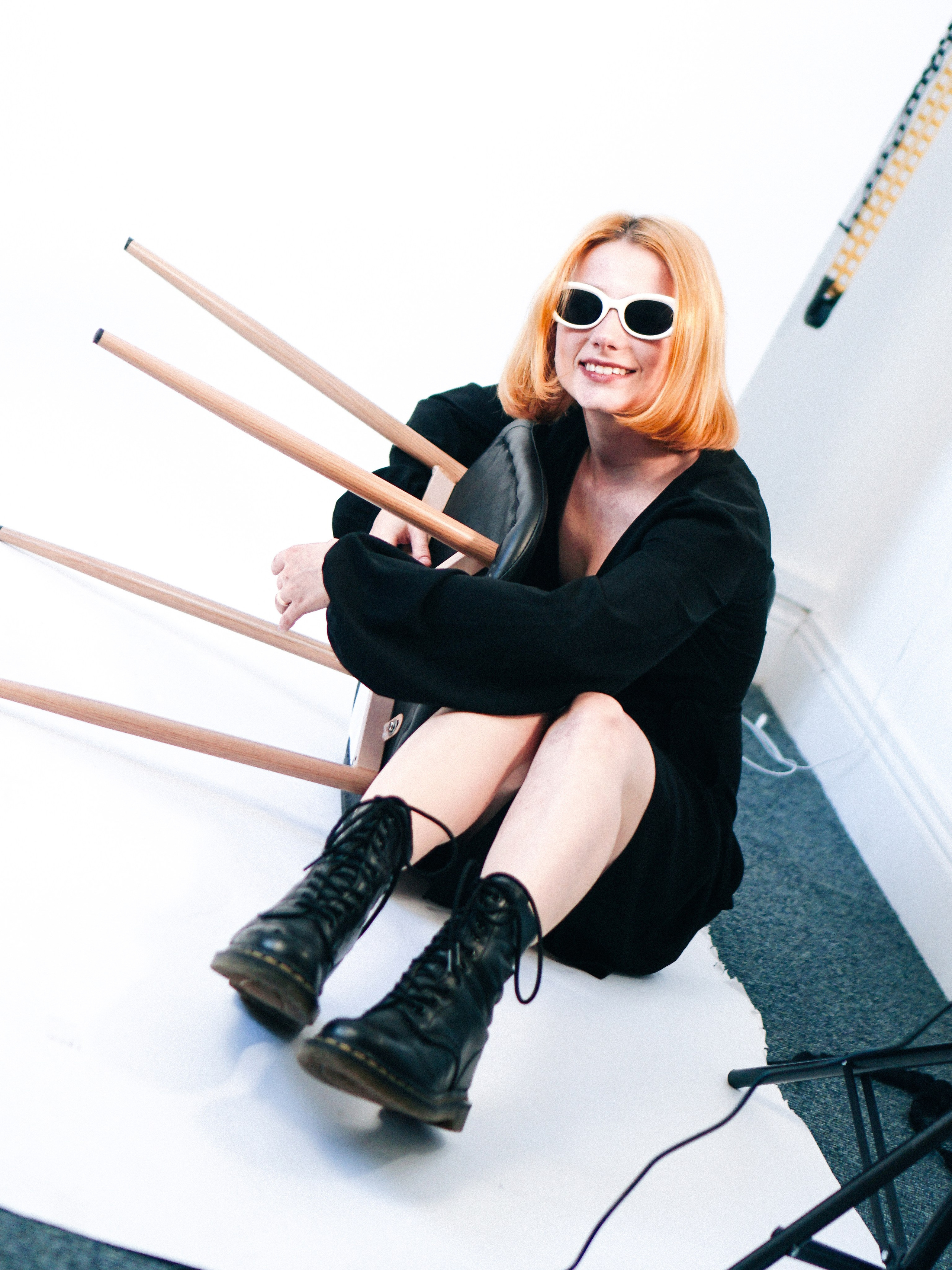 Redhead model in black dress and white sunglasses sitting playfully on the floor with an upside-down chair