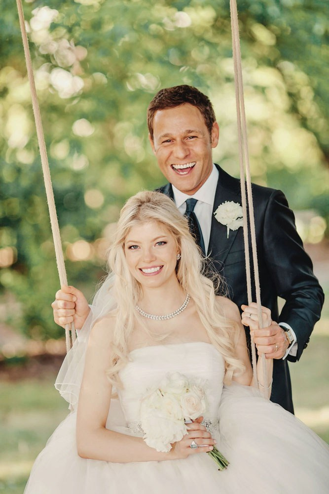 Elegant Swiss blonde bride on swing with bouquet, Italian groom caressing hand at summer wedding in Prague.