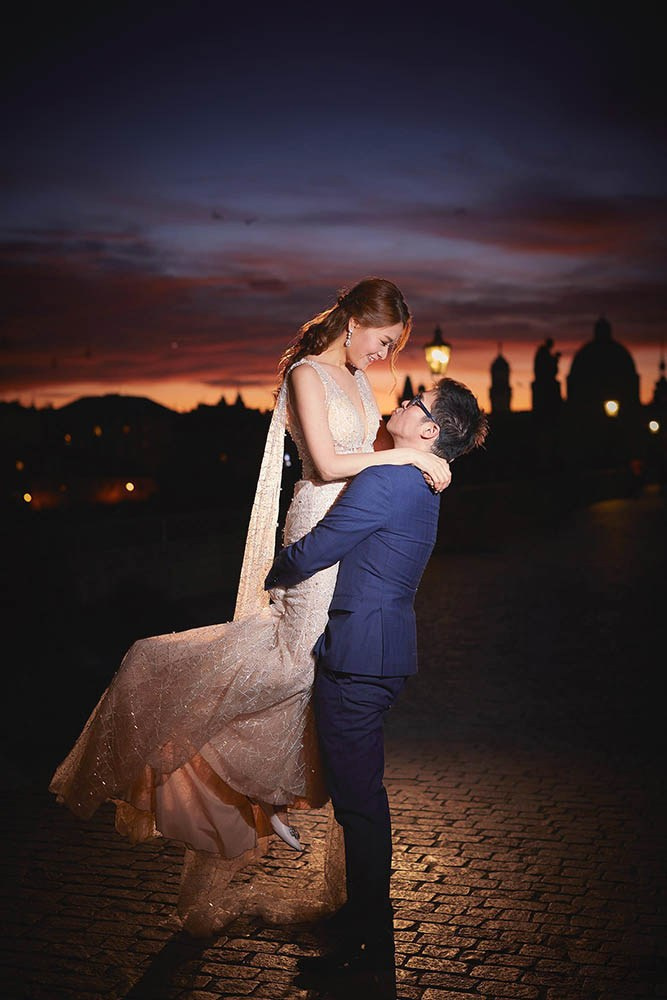 Groom lifting fiancée under gas lamps with flaring sky, Charles Bridge.