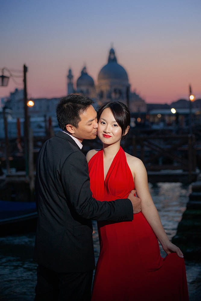 Thai woman in a red dress is kissed by her partner as they stand near the gondola in Venice at sunset.