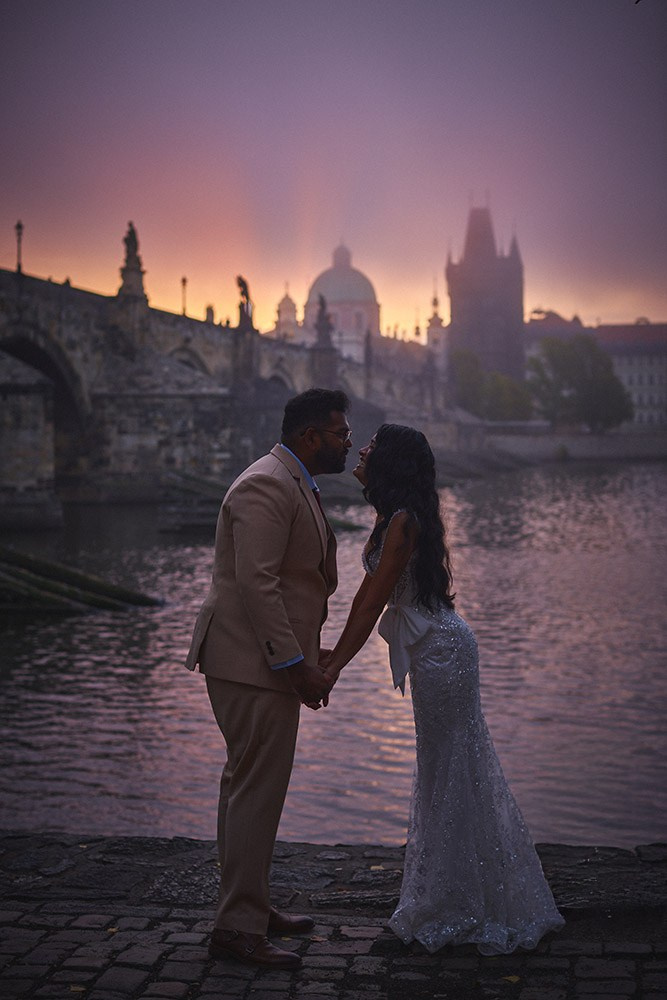 Newlyweds are nearly silhouetted against the rising sun above the Charles Bridge