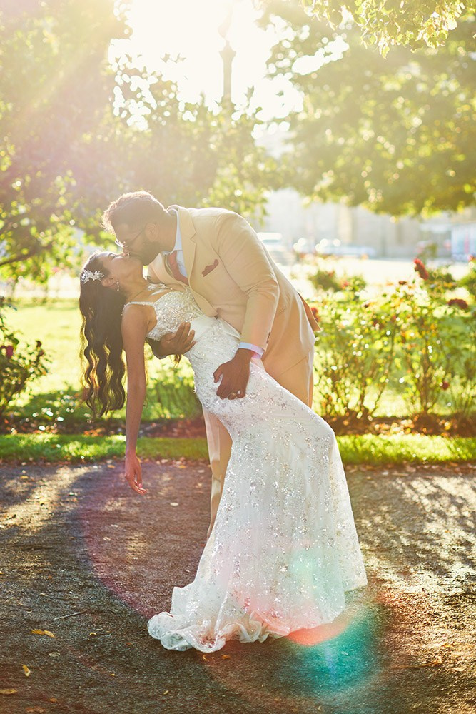 As sun flares vividly behing them, a bride is lovingly dipped and kissed by her groom near roses on the grounds of the Rudolfinum Symphony House