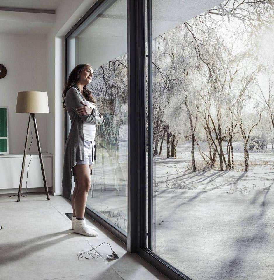 Девушка стоит в комнате рядом с панорамным окном и любуется зимним пейзажем в ясный солнечный день.