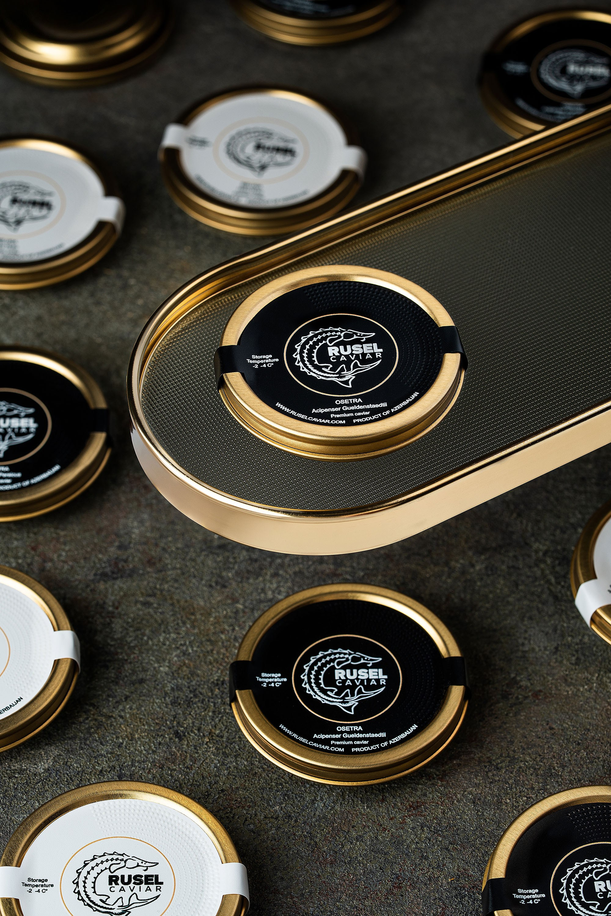 Tünd səth üzərində Rusel Caviar qapaqlarının düzümü, qızılı platforma üzərində əsas məhsul, yumşaq istiqamətli işıqlandırma ilə metal detalların vurğulandığı premium məhsul fotoqrafiyası.