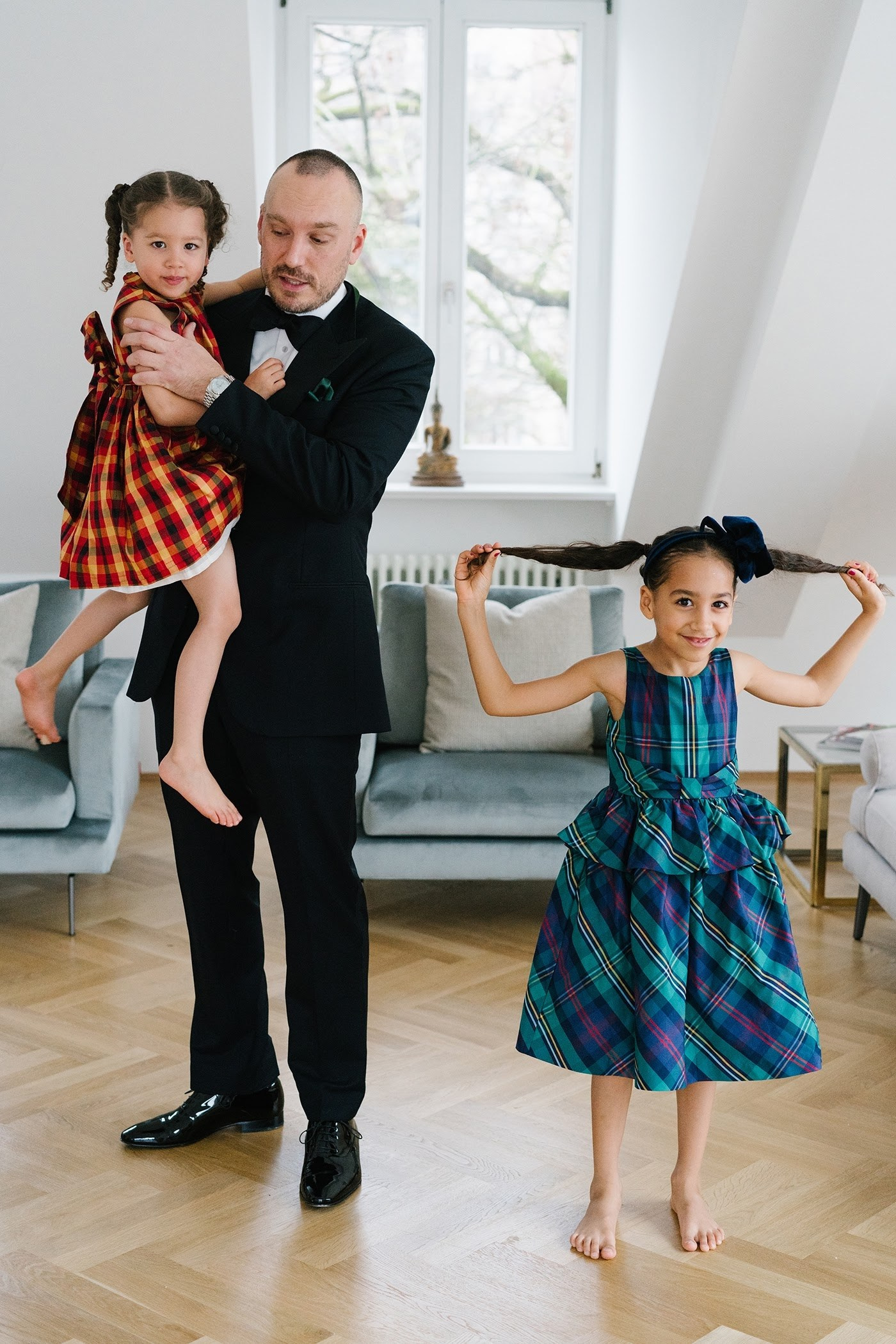 A father in a tuxedo holds his young daughter in a red dress while her older sister in a blue plaid dress plays with her hair in a bright Munich apartment during a cosy Christmas family photo shoot