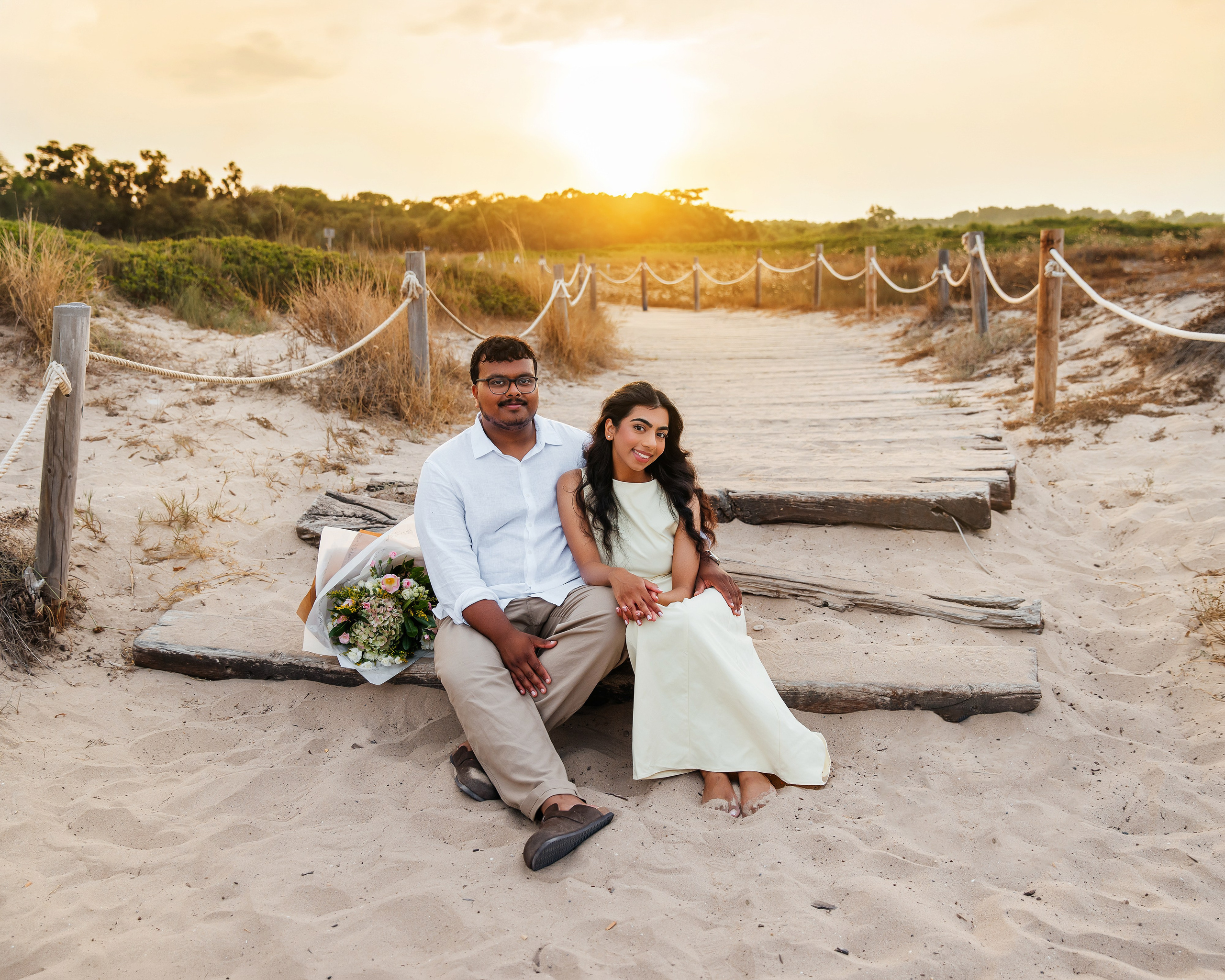 Couple portrait on wooden steps among sandy dunes, with a bouquet beside them during a proposal photoshoot.
