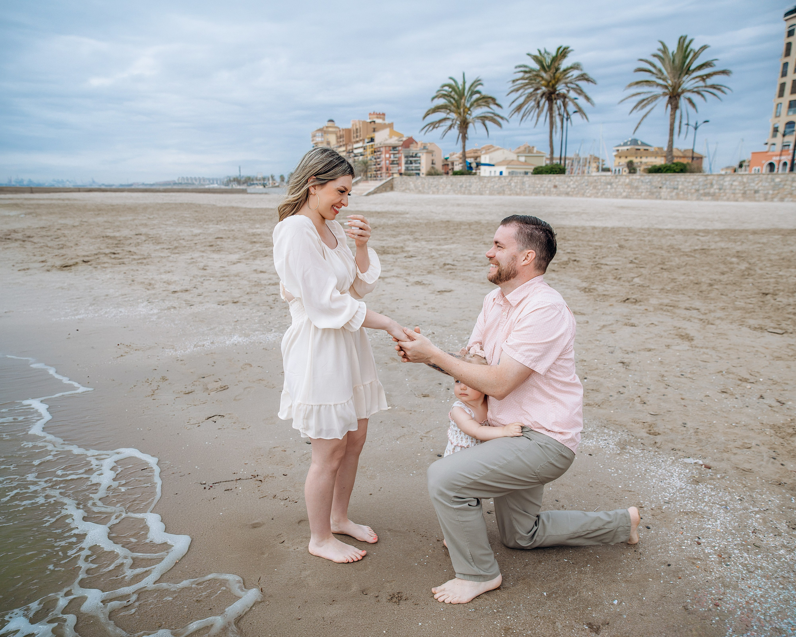 Man kneeling and holding his partner’s hand during a surprise proposal on Port Saplaya beach, with palm trees behind them.