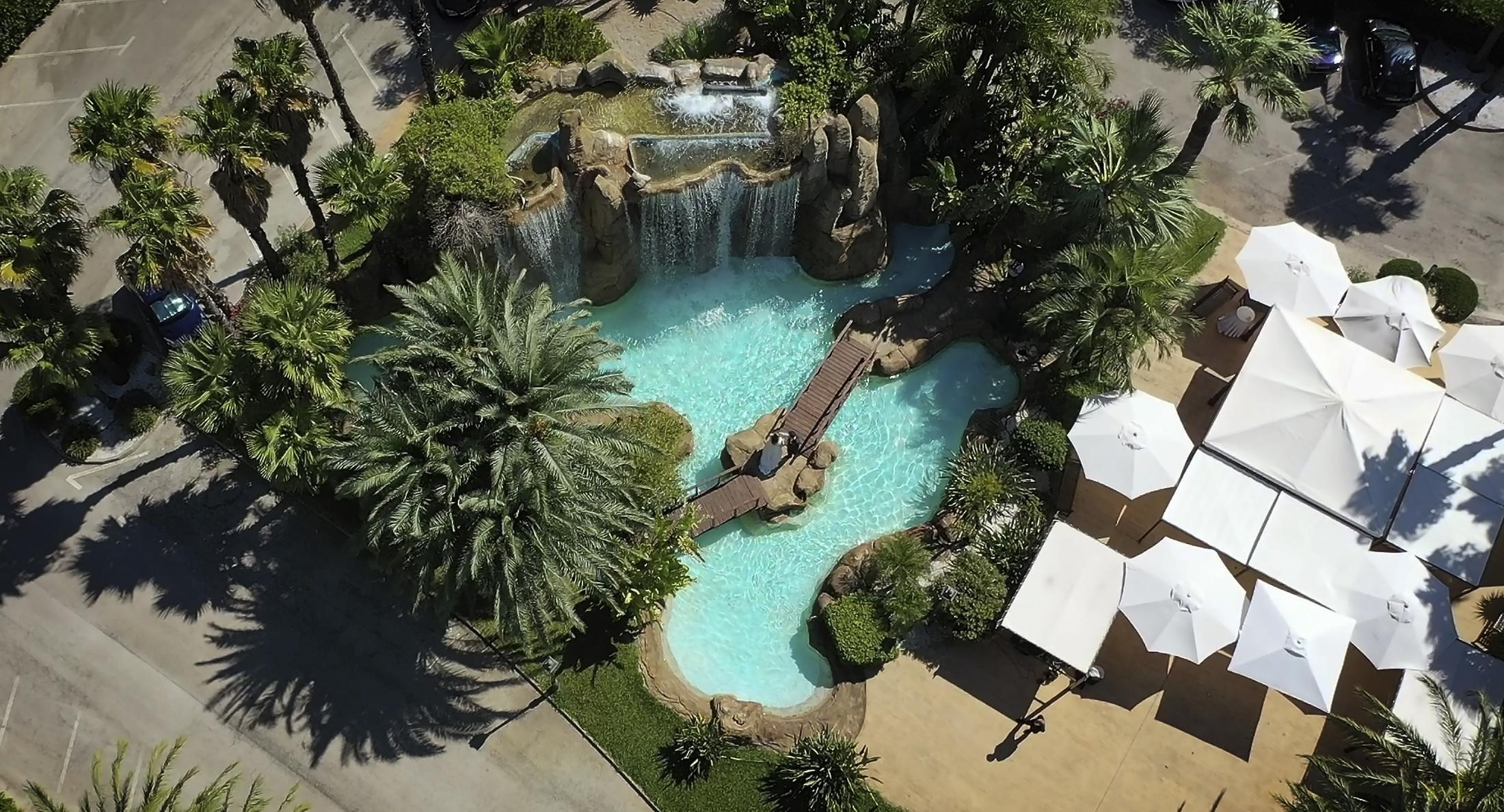 Aerial view of Jardines El Poblet’s turquoise pool, waterfall and wooden footbridge, surrounded by palms and white parasols.
