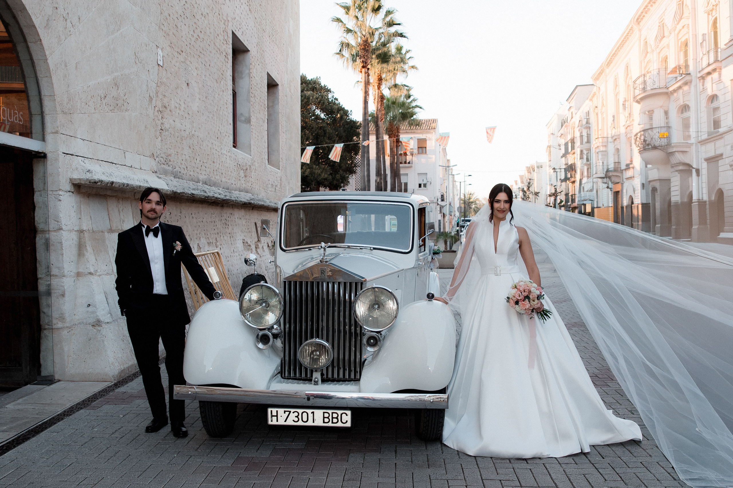 Bride and groom beside a vintage wedding car on a city street lined with historic buildings.