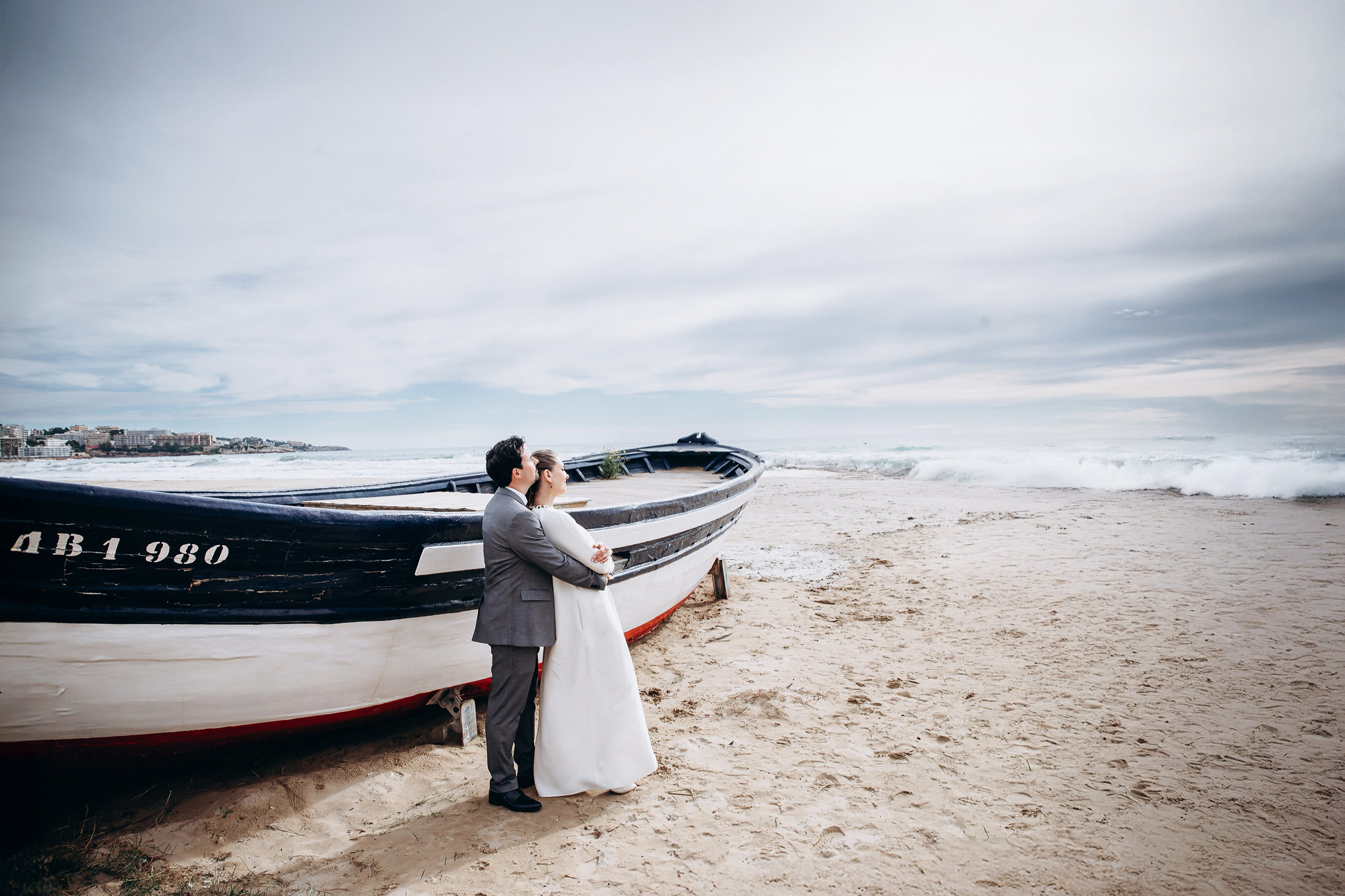 Bride and groom embracing beside a fishing boat on a sandy beach, with the sea and cloudy sky behind them.