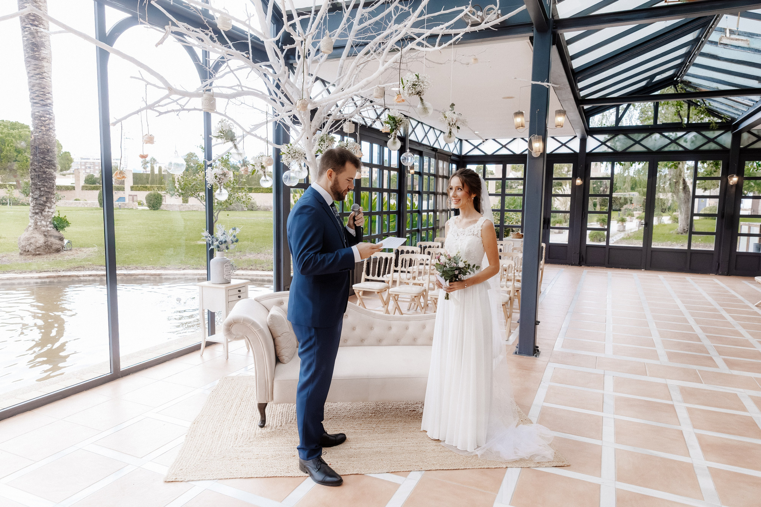 Bride and groom facing each other during a wedding ceremony in a glass pavilion overlooking the gardens.