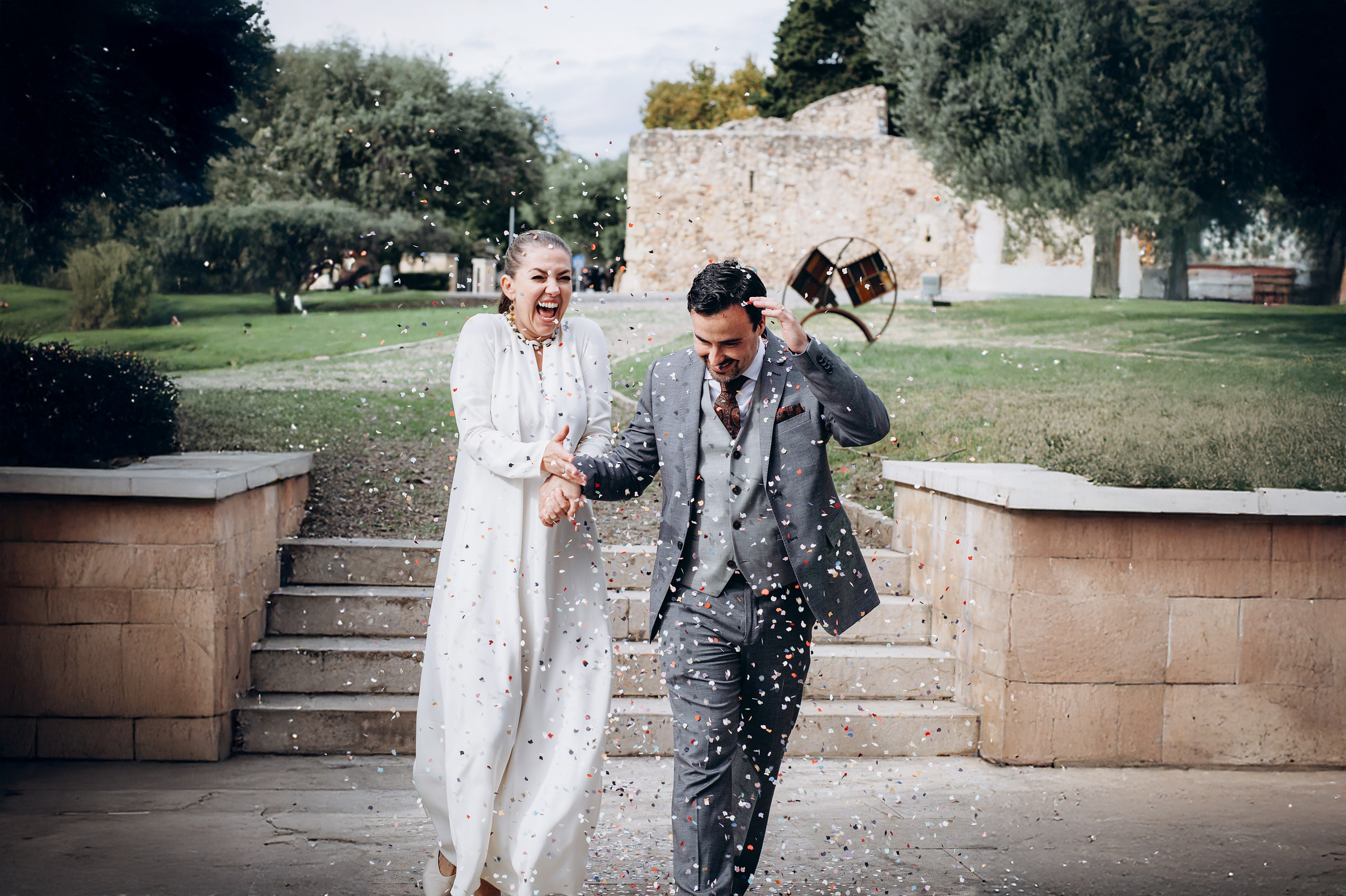 Newlyweds walking through falling confetti at the foot of stone garden steps.