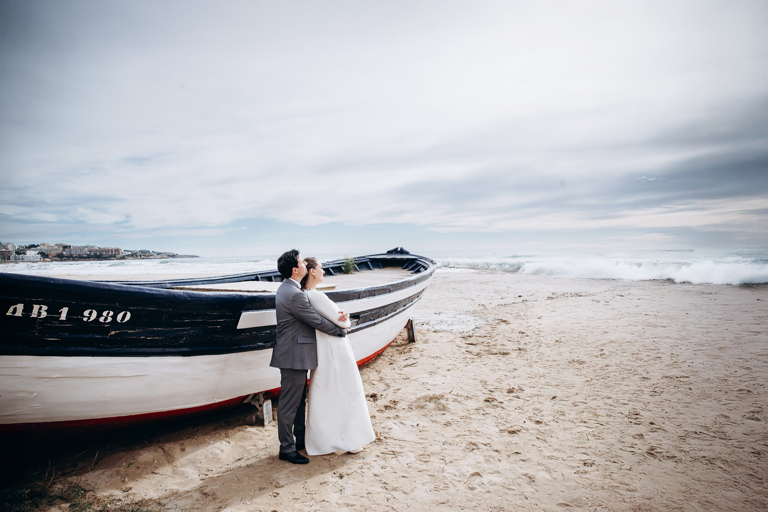 Свадебная фотосессия в Малаге, Испания, запечатлевшая объятия жениха и невесты на песчаном пляже рядом с традиционными рыбацкими лодками у моря, с мягким естественным светом и спокойной средиземноморской атмосферой — идеально для блог-статей о свадебной фотографии в Малаге, пляжных свадебных фотосессиях на юге Испании, свадьбах за границей, романтических прибрежных свадебных портретах и профессиональной свадебной фотографии по всей Испании.