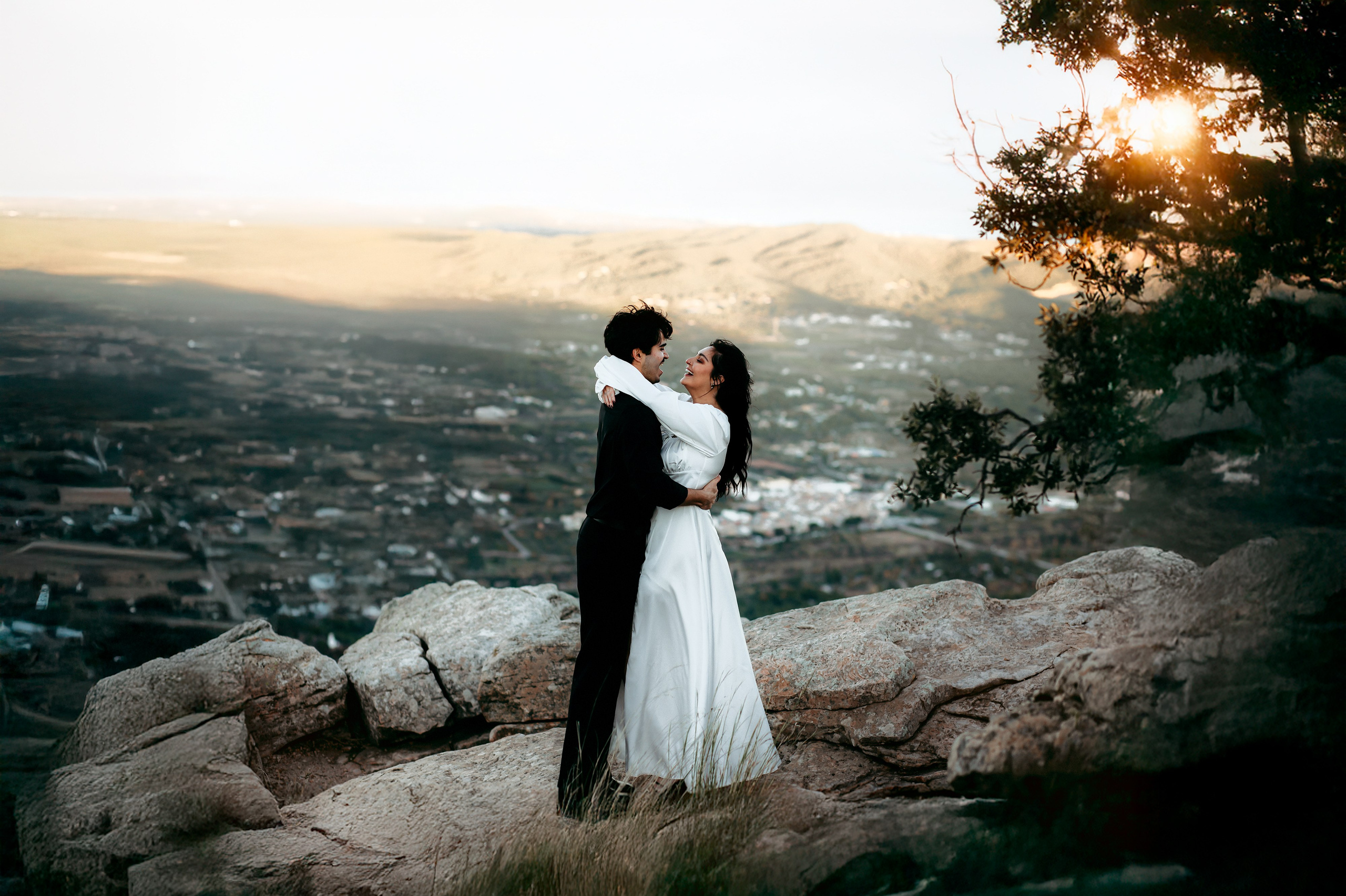 Couple embracing on a rocky mountain viewpoint near Valencia, with sunlight filtering through the trees.