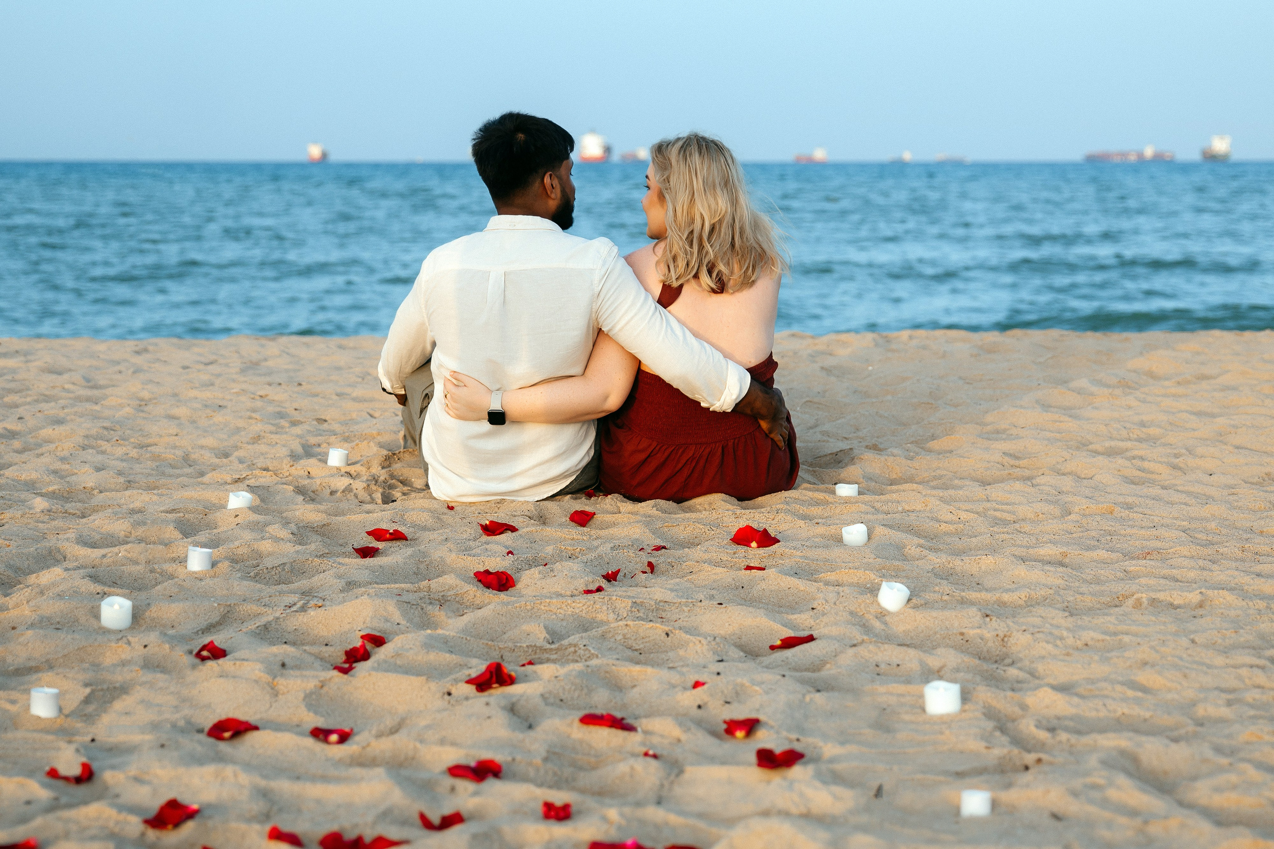 Couple sitting arm in arm facing the sea, with rose petals and candles arranged on the sand for a beach proposal in Valencia.
