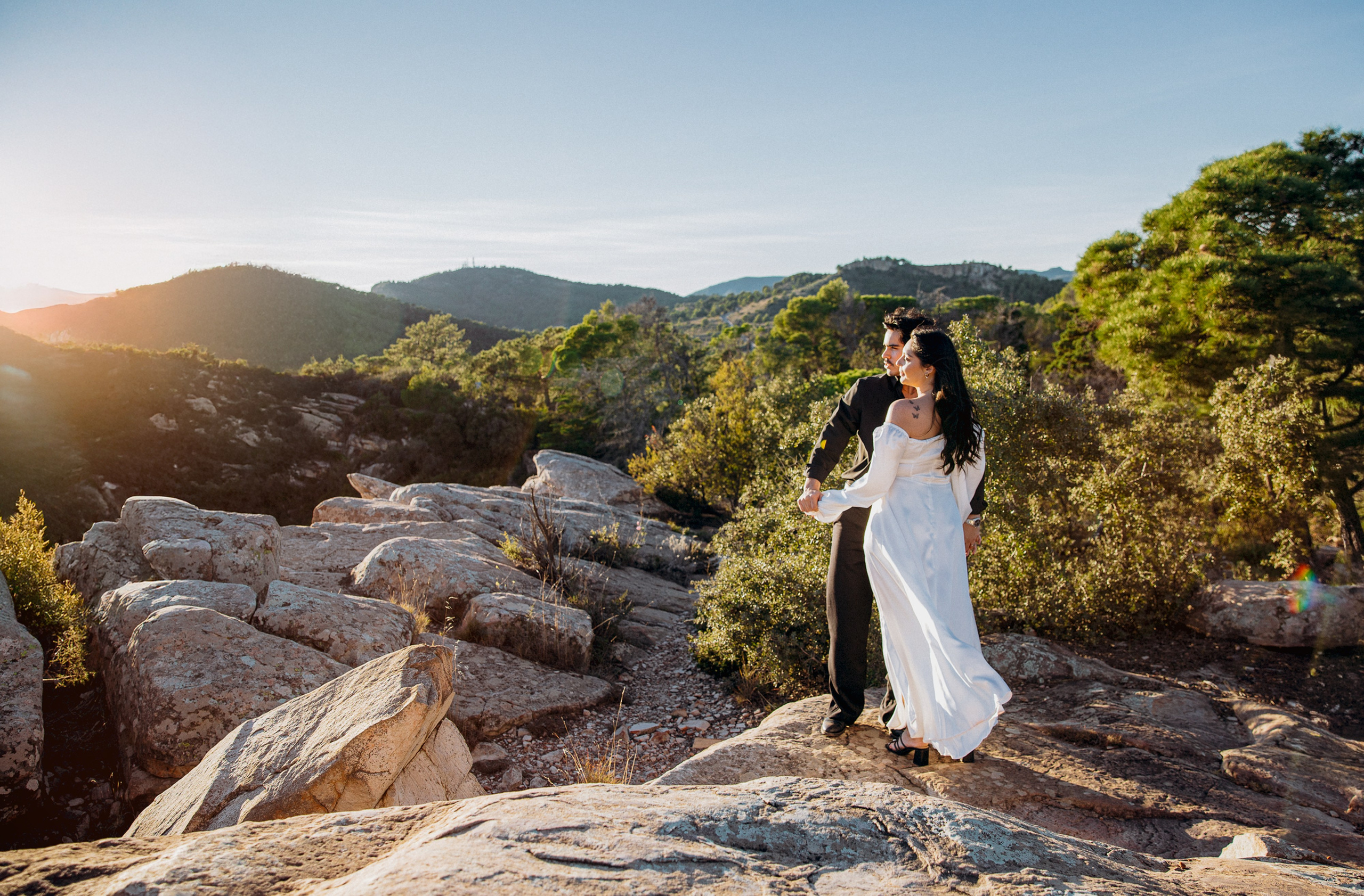 Свадебная фотосессия в Гранаде, Испания, где жених и невеста идут вместе по живописным каменистым холмам в золотой час, в окружении пышной зелени и тёплого света заката, передавая романтичный и кинематографичный момент — идеально для блог-статей о свадебной фотографии в Испании, свадебных фотосессиях в Гранаде, свадьбах за границей, естественных свадебных портретах и профессиональной свадебной фотографии по всей Испании.