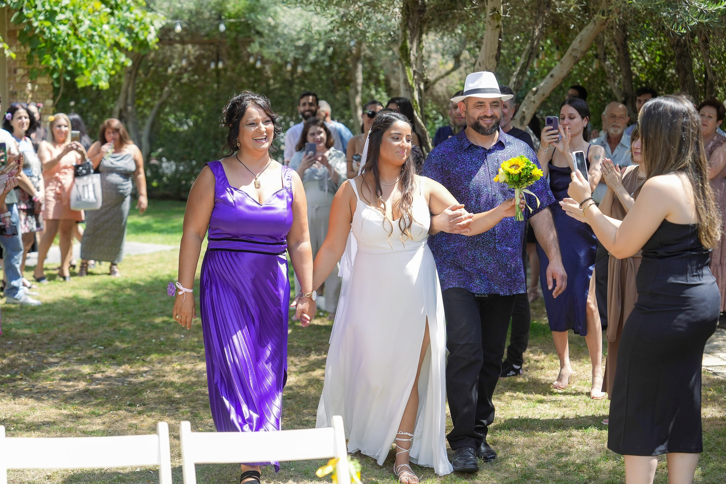 A bride walks down the aisle with her family during an outdoor wedding ceremony