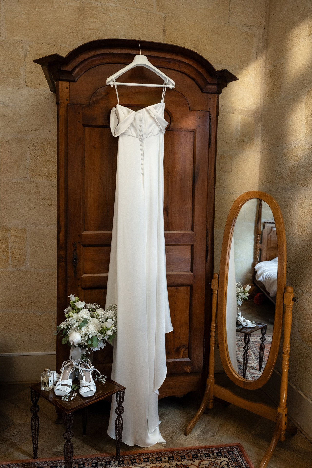 Robe de mariée suspendue dans une chambre de château en France, avec bouquet et chaussures préparés pour la cérémonie.