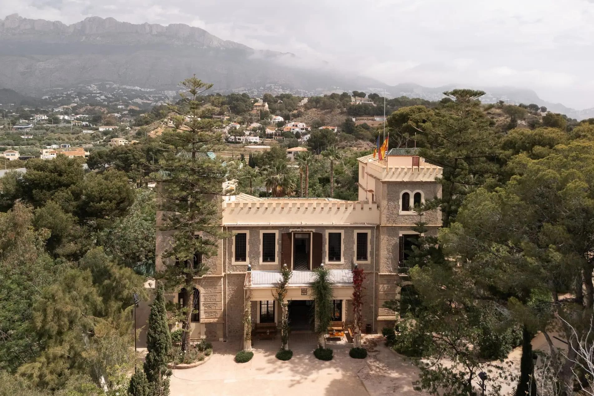 Elevated view of Finca Marqués de Montemolar’s towered villa, surrounded by trees with mountains in the distance.