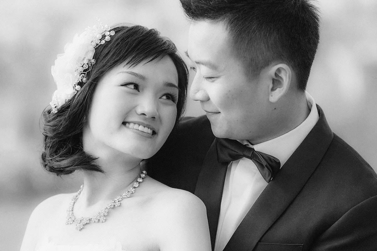 A smiling Hong Kong bride is embraced from behind by her bow-tie-wearing partner during their Prague pre-wedding.