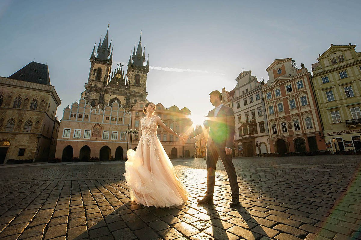 A radiant Singapore woman lovingly holds her partner's hand as the sun rises behind them above the spires of Tyn Church in an empty Old Town Square in Prague.