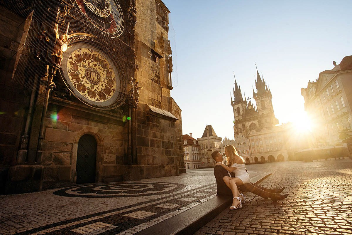 Blonde-haired woman smiling at her partner while sitting in his lap underneath the Astronomical Clock in Prague as the sun flares behind them in an empty Old Town Square at sunrise.