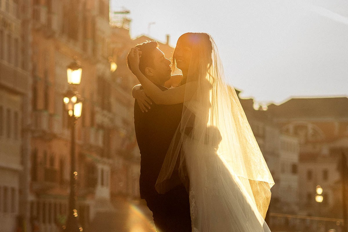A bride smiles as she plays with her groom's hair as he holds her aloft, surrounded by the warmth of early morning sunlight in Venice, Italy - Kurt Vinion Photography