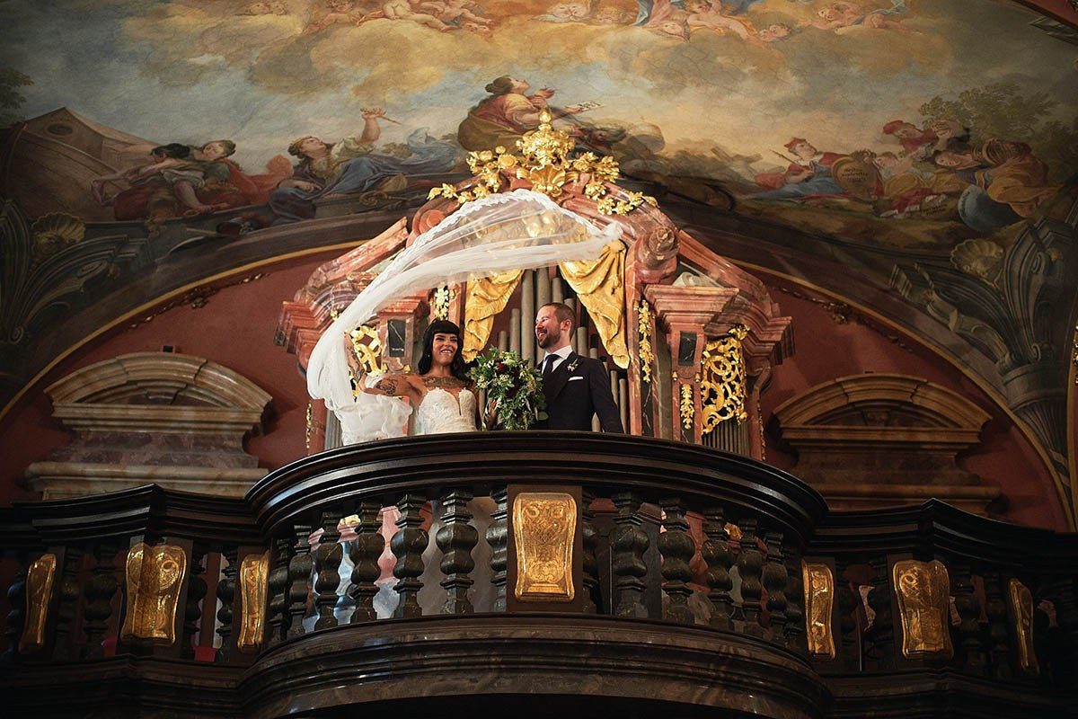 Radiant dark-haired tattooed-bride twirls her veil above her groom's head as he laughs, as they stand near the historic Mirror Chapel organ, as seen from below.