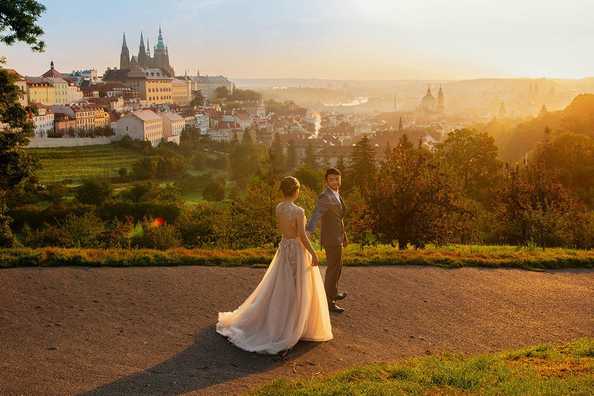 Young newlyweds from Singapore walk above the vineyards as the sun breaks above the skyline, illuminating the spires of St. Vitus and Prague