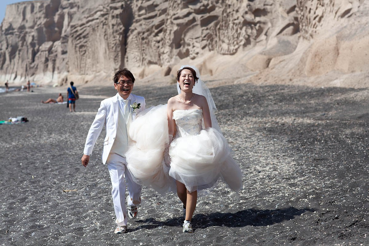 Junji and Nao walk hand in hand on the black sand beach in Santorini.