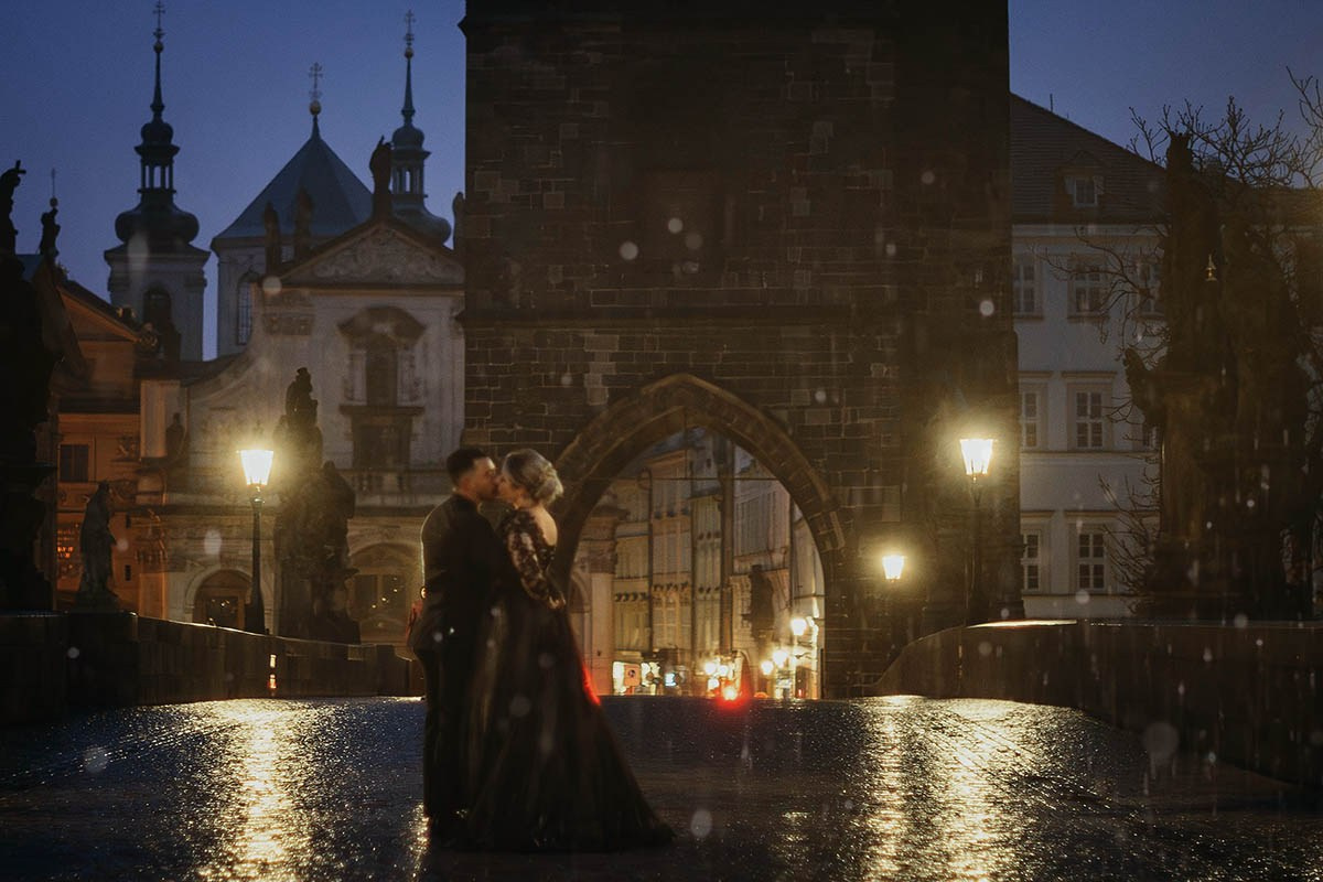 Couple in black, embracing atop the nighttime landscape of the Charles Bridge as it rains.