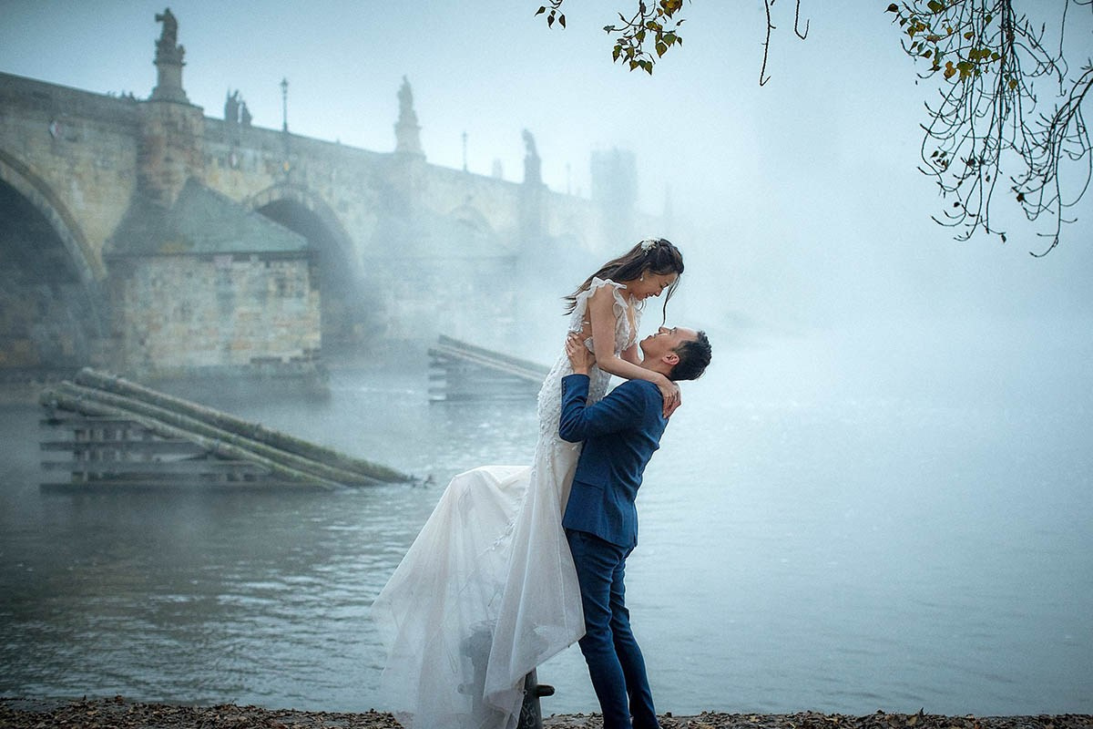 A radiant woman from Singapore lovingly holds her partner's hand as the sun rises behind them above the spires of Tyn Church in an empty Old Town Square in Prague.