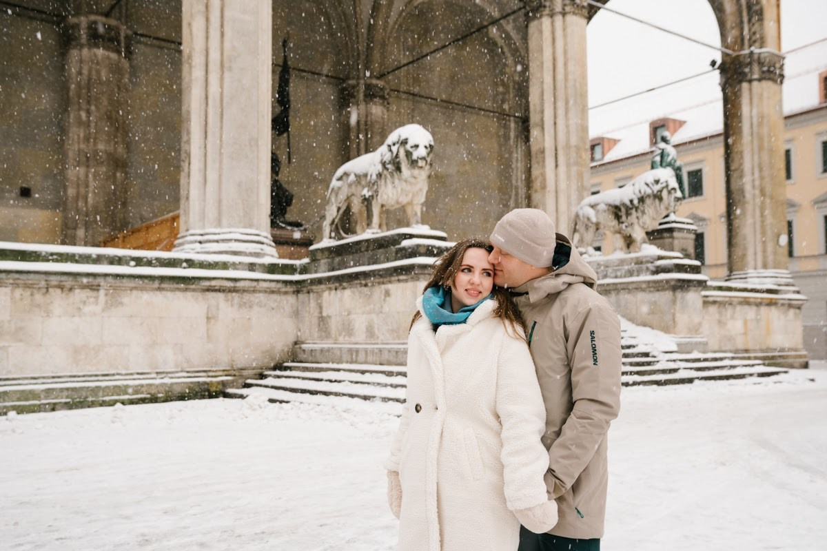 A romantic couple cuddling in warm winter coats under falling snow in front of historic statues in Munich city center during a Christmas photosession