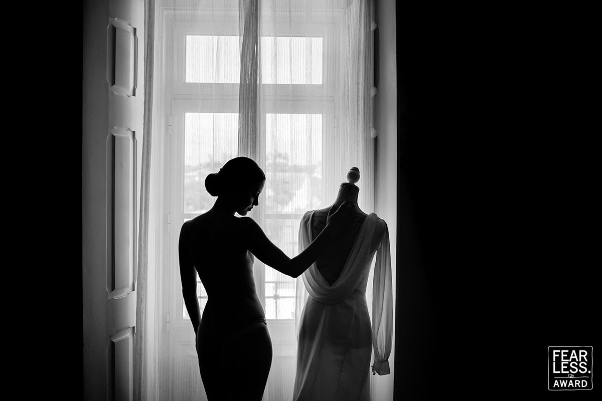 Bride in silhouette beside her wedding dress in front of a bright window