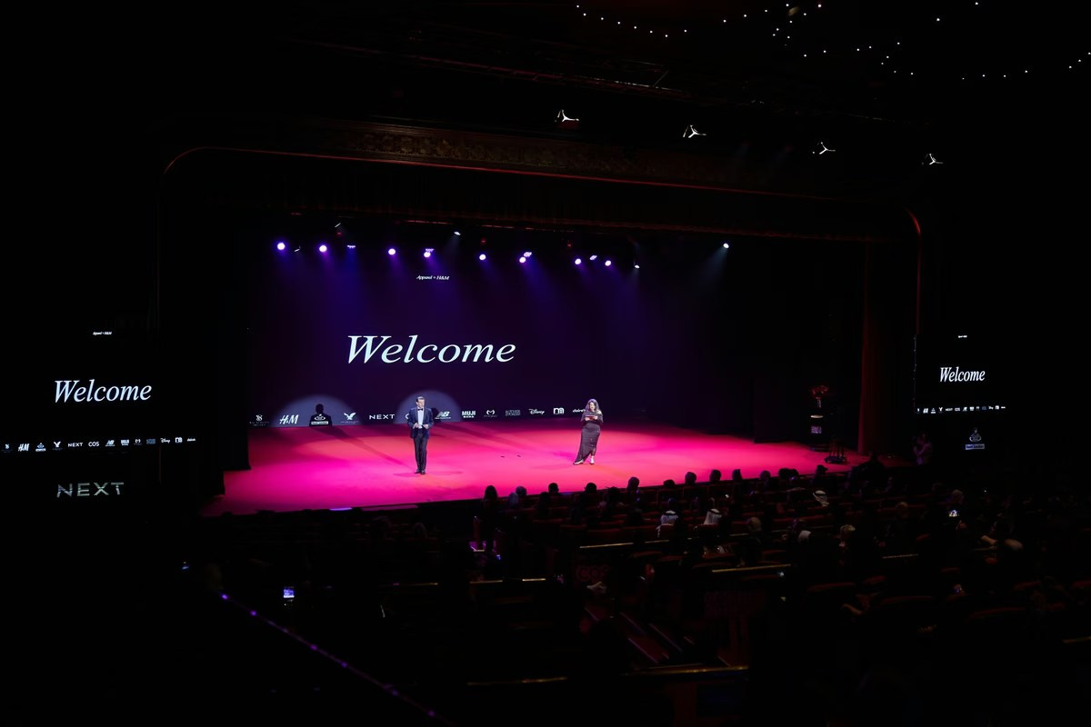 Wide view of a corporate conference stage with two presenters, branded screens and an audience in Dubai
