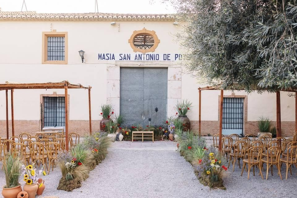 Outdoor wedding ceremony setup at Masía de San Antonio de Poyo, with wooden chairs and a flower-lined gravel aisle.