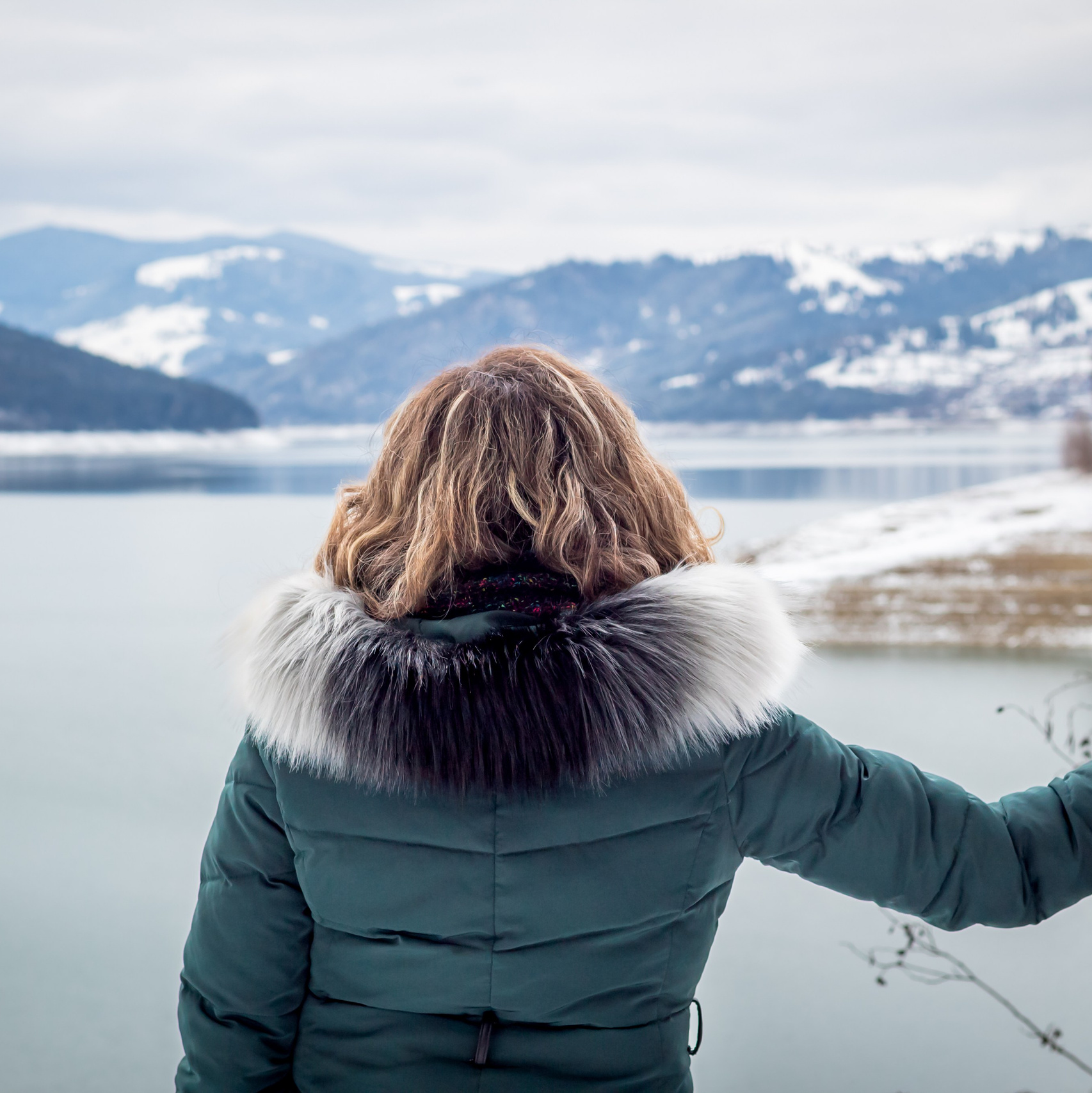 Woman in winter coat looking out over a frozen lake with snow-covered mountains in the distance
