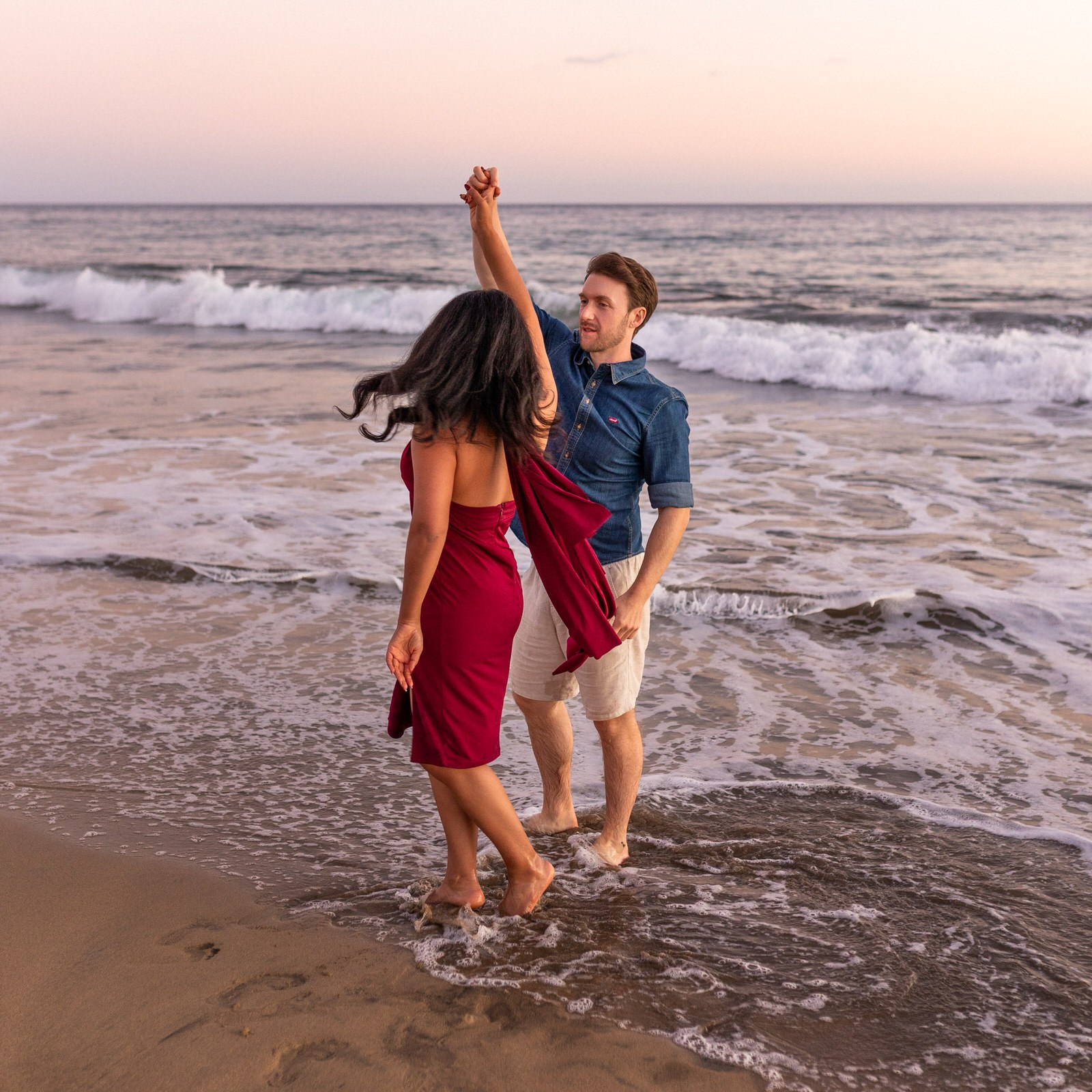 A couple dances barefoot on the wet sand at the beach near the shoreline during sunset, with gentle waves in the background on Meloneras
