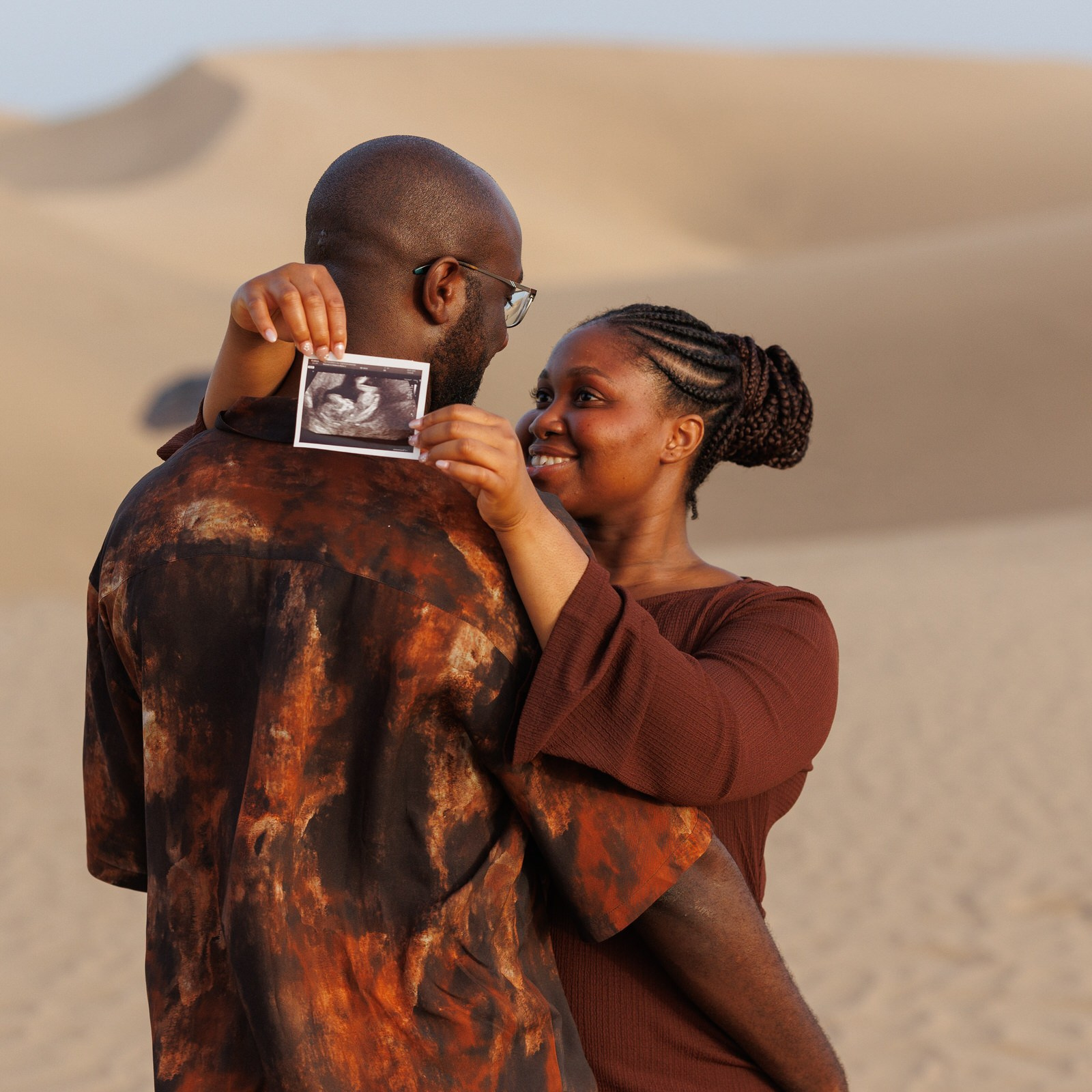 Maspalomas Gran Canaria A couple stands in a desert, embracing while one person holds up a sonogram image, both looking at each other and smiling.