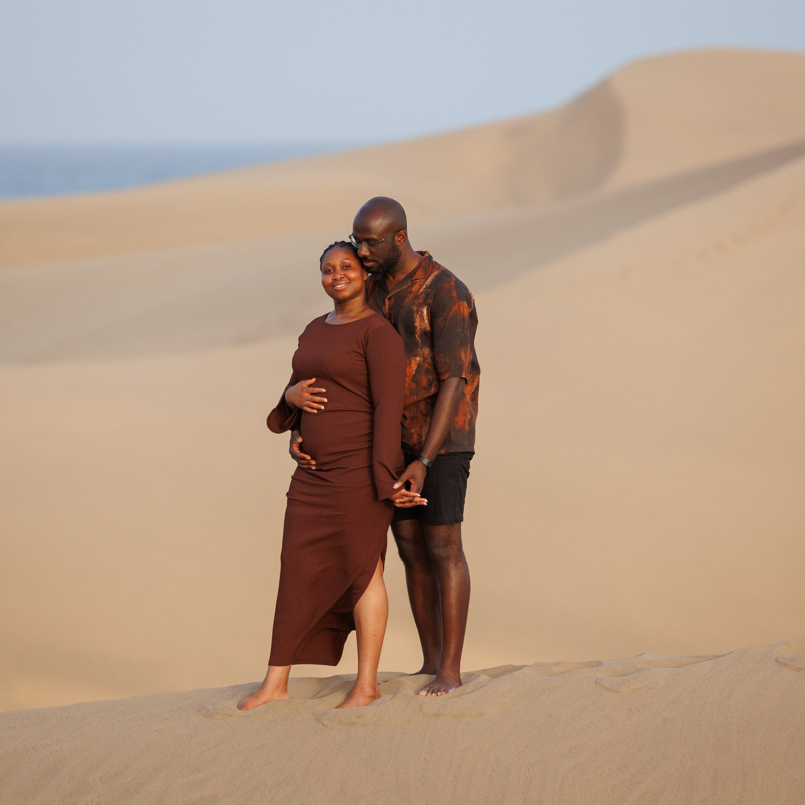 Maspalomas Gran Canaria A pregnant woman in a brown dress stands on sand dunes, smiling, while a man stands behind her holding her hand.