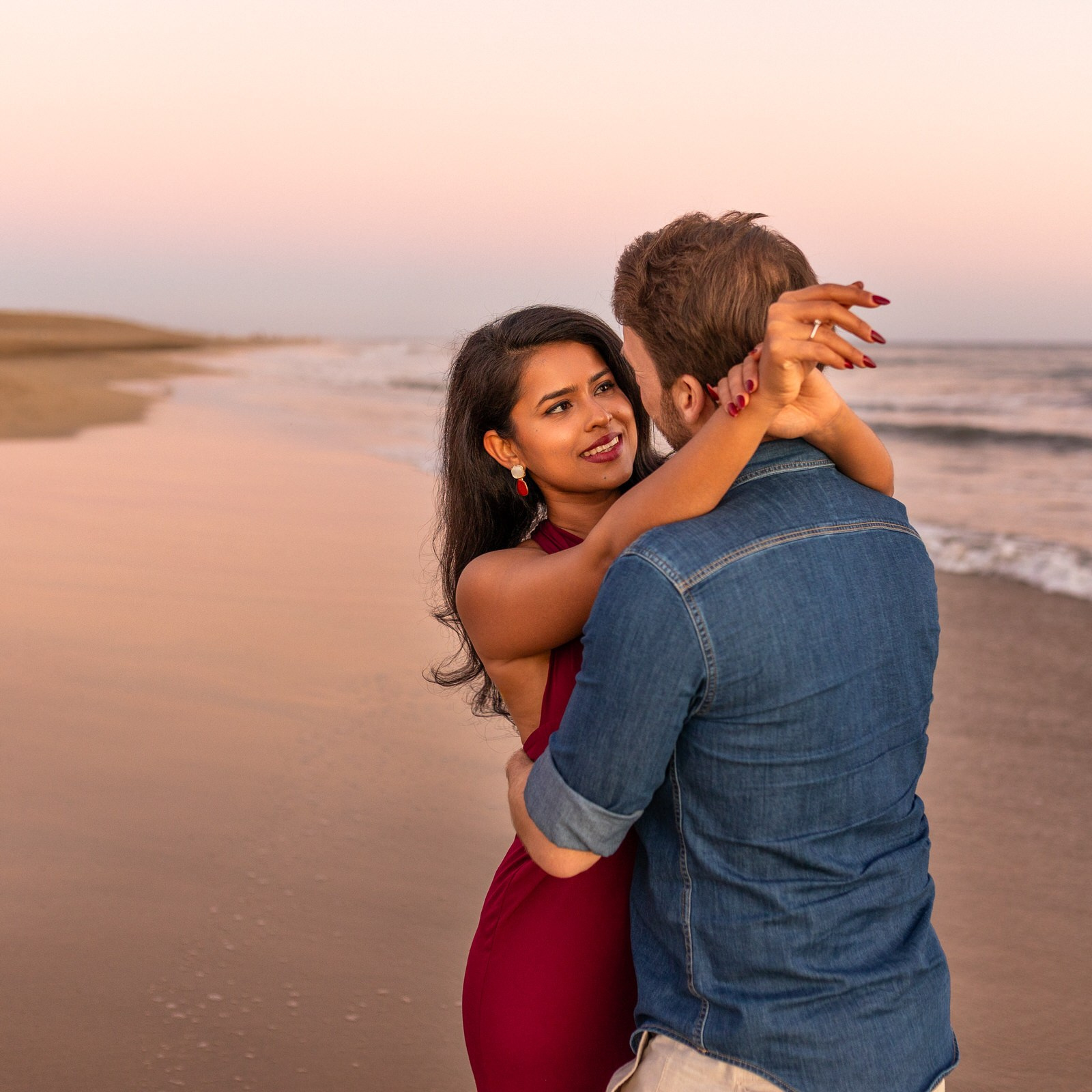 A couple stands embracing on a sandy beach at sunset, with gentle waves and a pastel sky in the background.