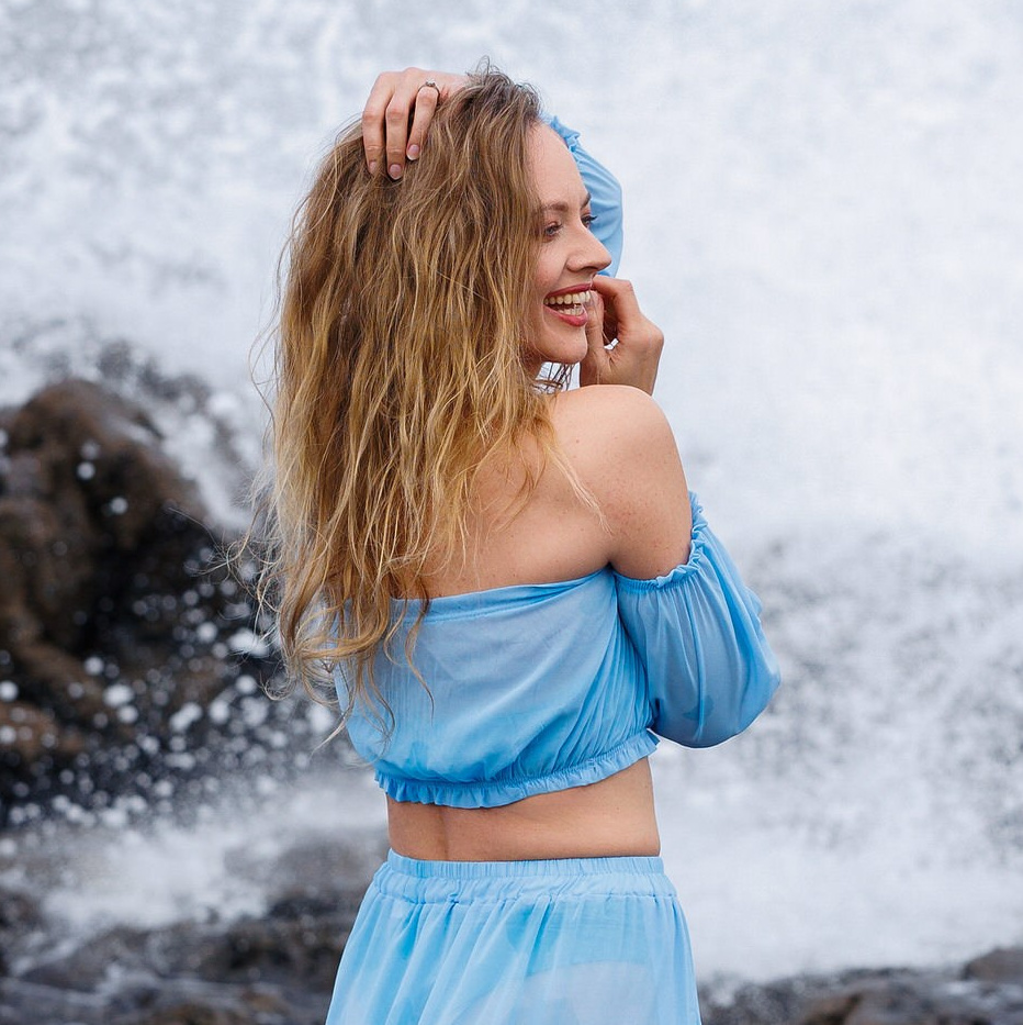 Smiling woman with windblown blonde hair in blue, ocean spray behind, Gran Canaria's volcanic coast