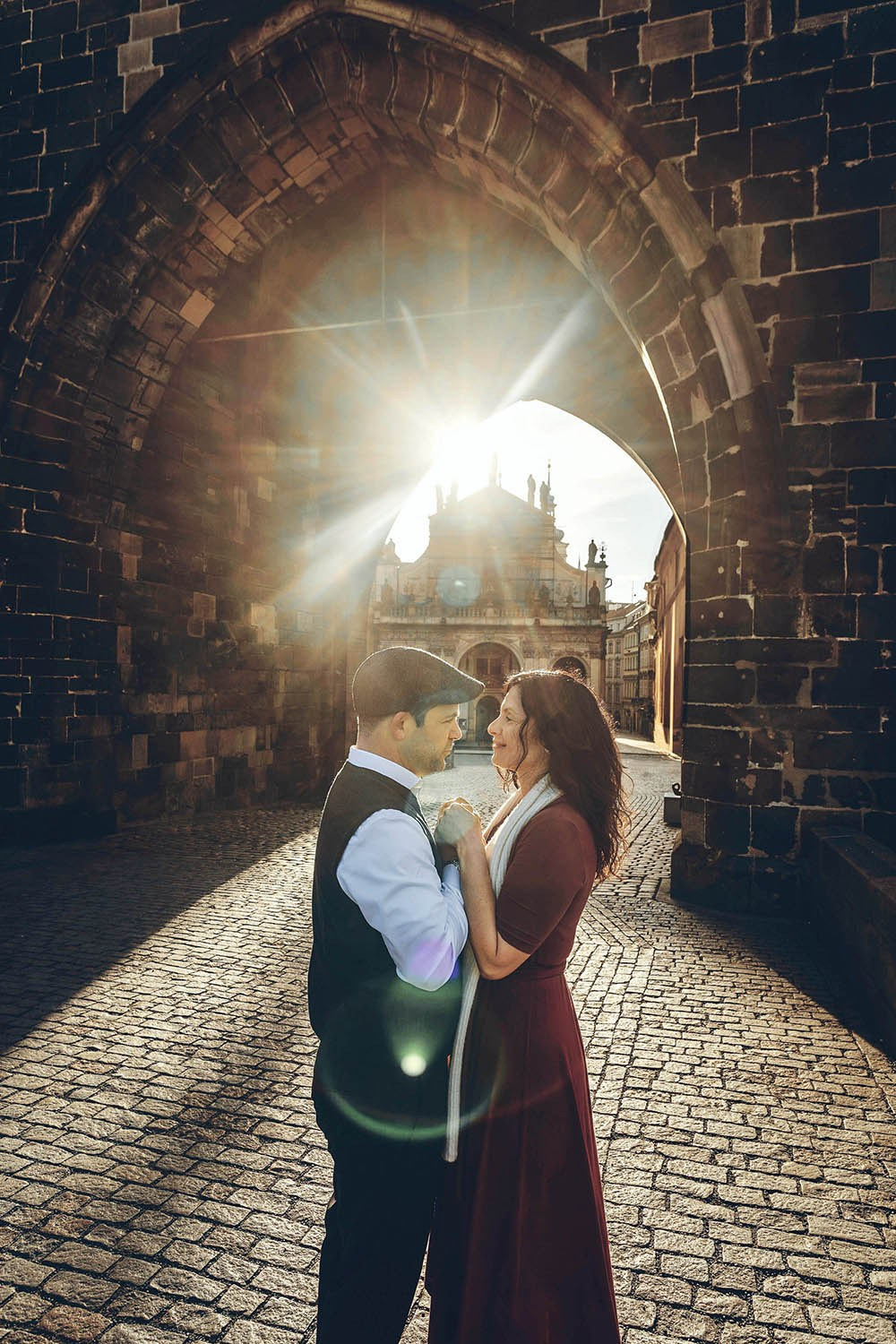 Couple standing beneath the Gothic archway of Prague's Old Town Bridge Tower as sunrise light creates a cinematic lens flare effect