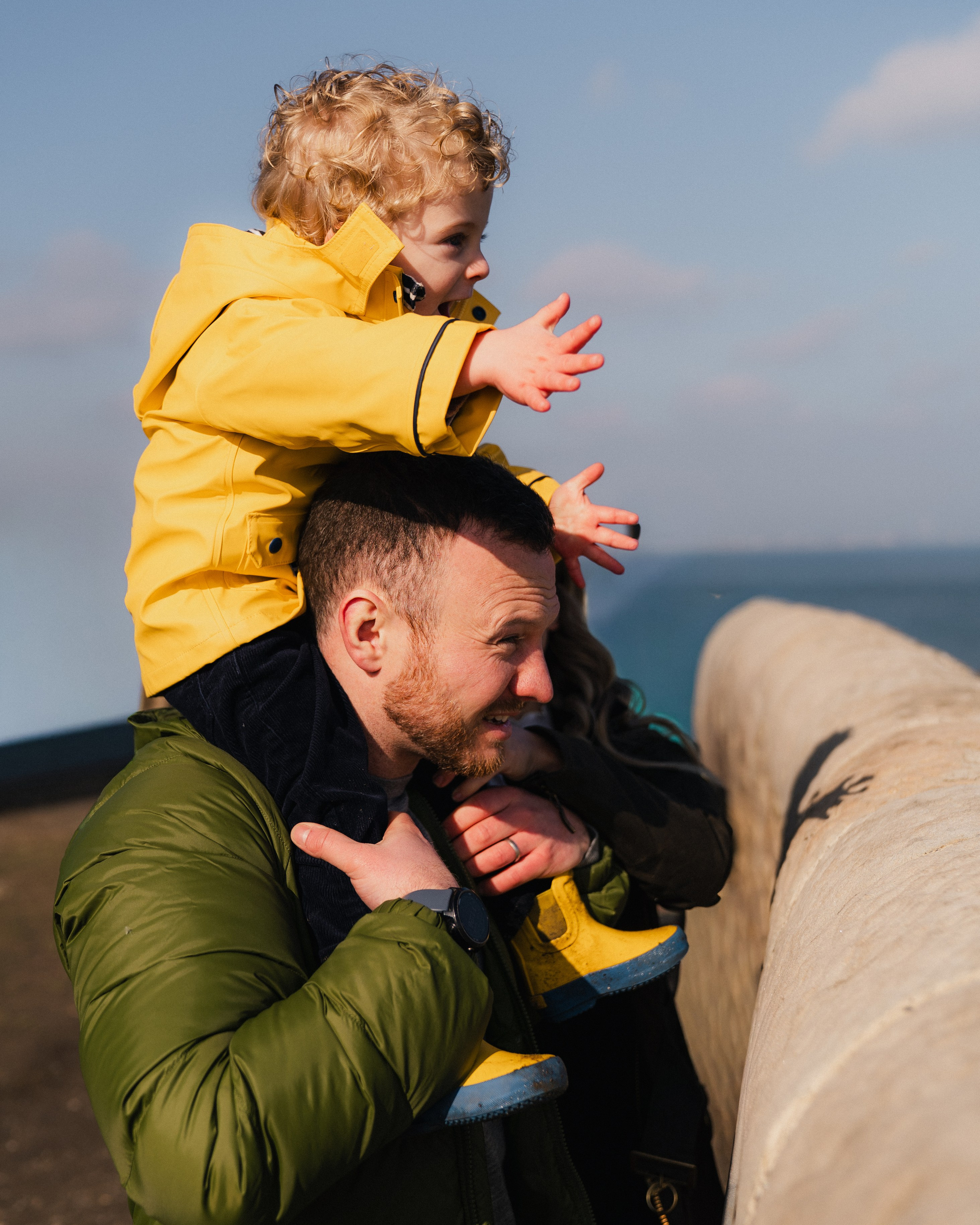 Child sitting on father’s shoulders during a family photoshoot by the sea in Whitley Bay, joyful and playful spring moment