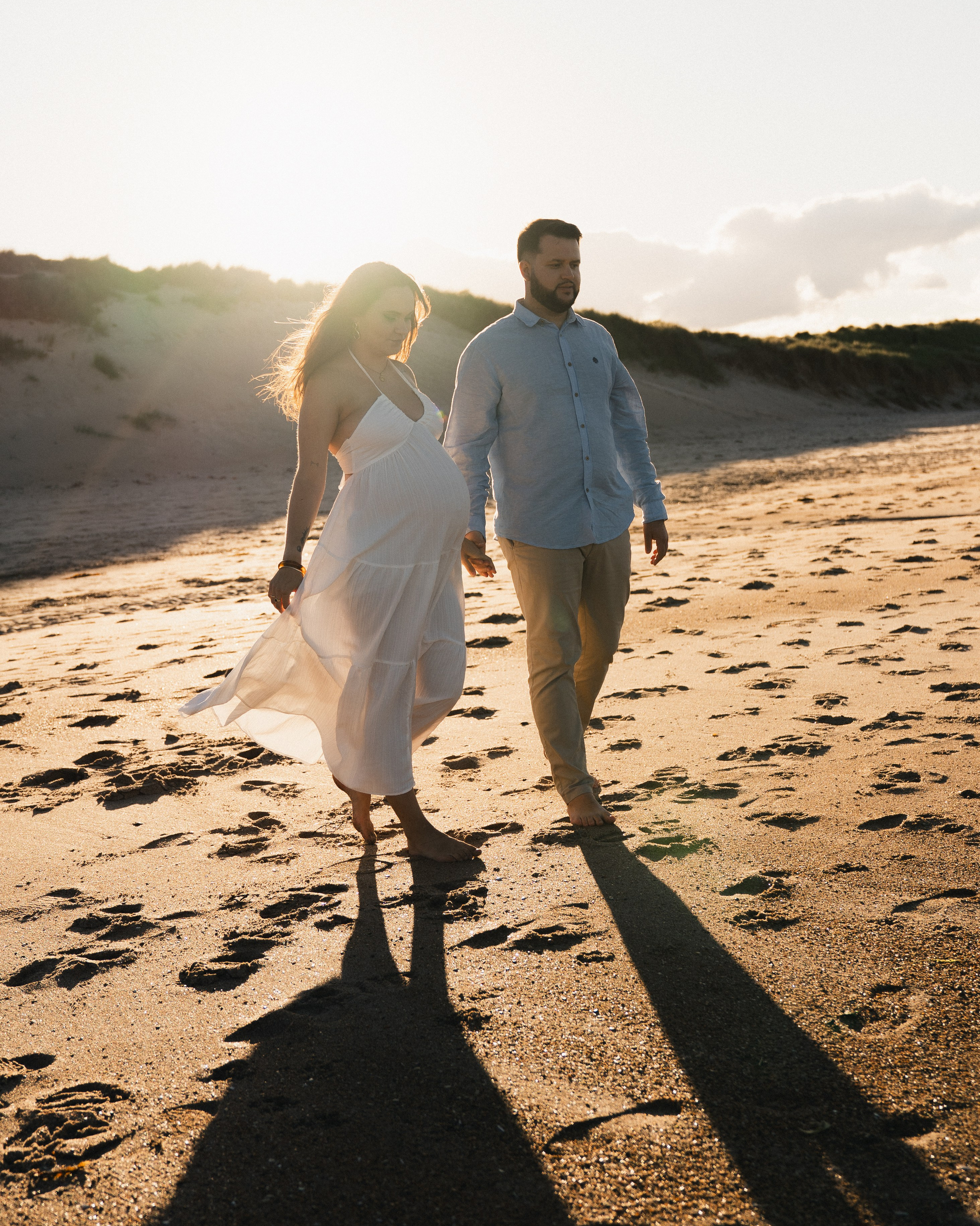 Pregnant couple walking hand in hand along Druridge Bay beach during a golden hour maternity photoshoot, with warm sunset light and long shadows.